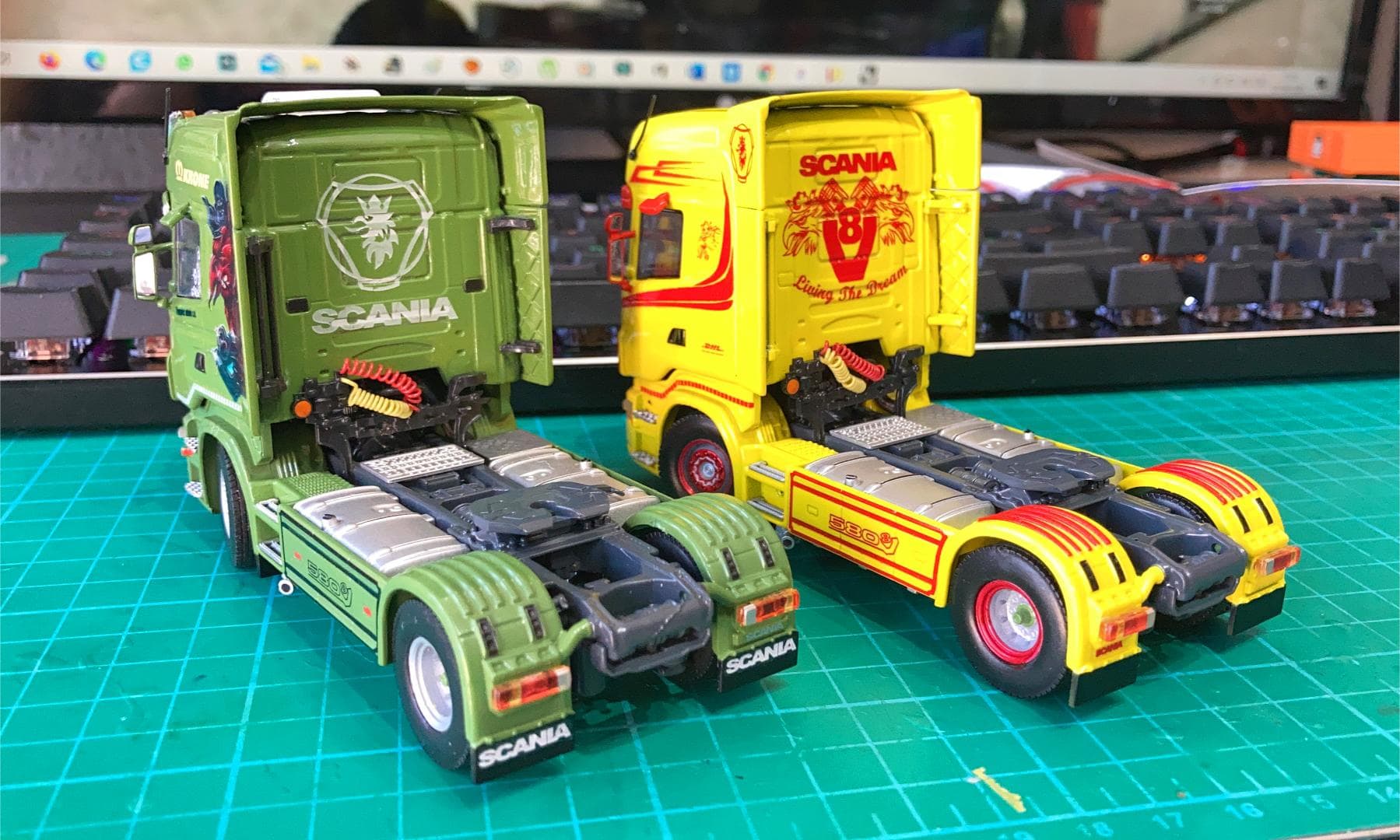 Dos modelos coloridos de camiones Scania se abren camino a través de la alfombra de corte verde, simulando viajeros del Camino superando obstáculos, con un teclado y un monitor como fondo.