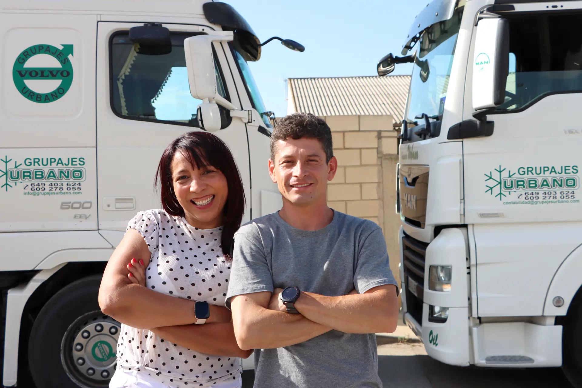 Dos personas sonrientes, de brazos cruzados, de pie delante de dos camiones con el logo de "Grupajes Urbano". Entre ellos, la reconocida camionera Luzilene Gomes, que encarna el espíritu de una amazona moderna, marcando con seguridad el camino en la logística.