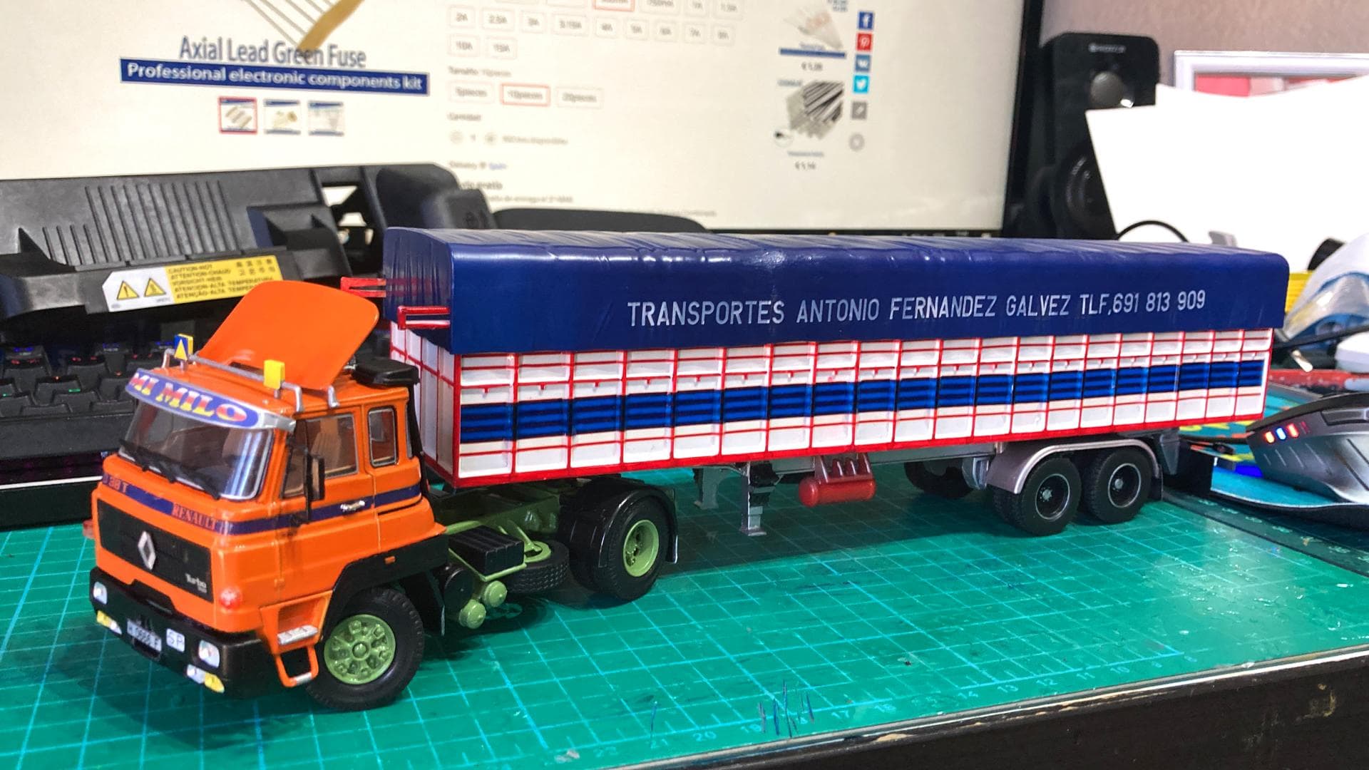 Camión modelo "Transportes Antonio Fernández Galvez" sobre remolque, navegando a través de un escritorio lleno de componentes electrónicos, como si estuviera superando obstáculos en la carretera.