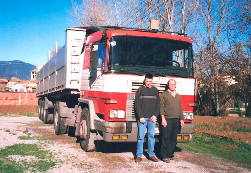 Dos hombres parados frente a un gran camión rojo TransMoradell estacionado en un camino rural con árboles al fondo.