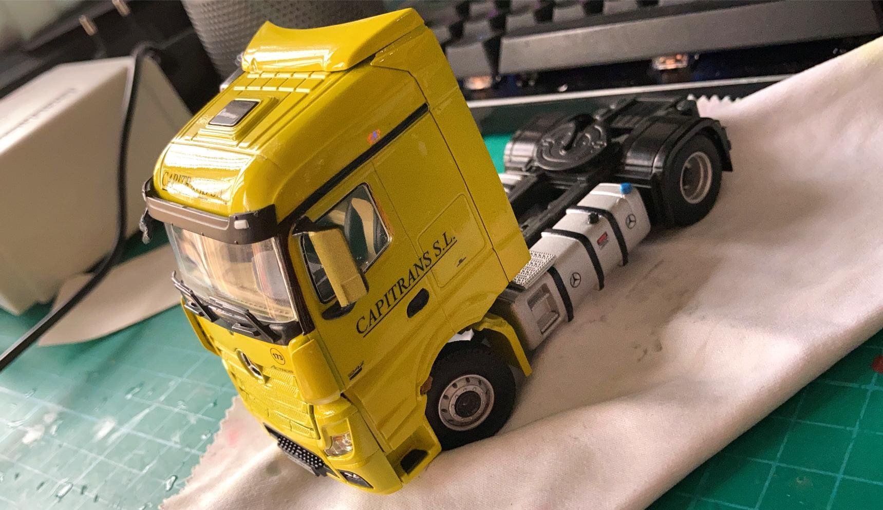 Maqueta de camión de librea amarilla con la leyenda "Capitrans S.L." en el lateral, expuesta sobre una tela sobre un tapete de corte verde, que recuerda el viaje de Antonio Fernández sorteando obstáculos a lo largo del camino.
