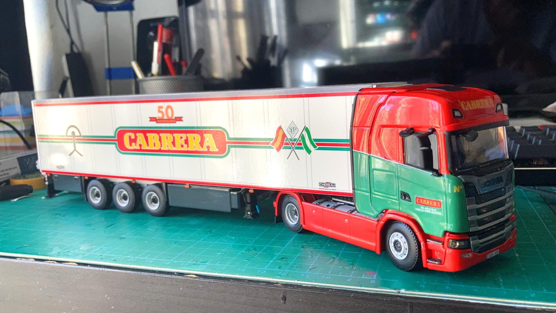 Un modelo detallado de un camión rojo y verde con el logotipo "Cabrera" en el remolque, exhibido en un banco de trabajo, captura el espíritu del viaje de Antonio Fernández por innumerables caminos, superando obstáculos en cada milla.