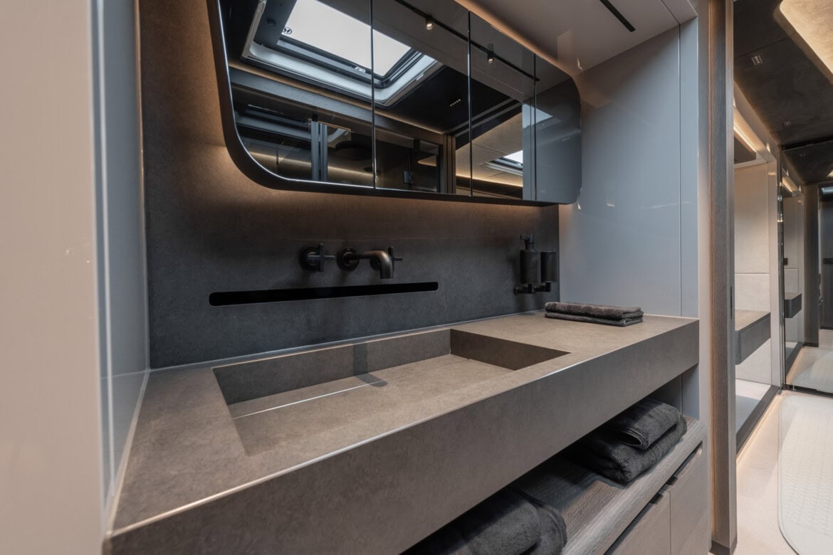 Baño moderno con encimera elegante, lavabo incorporado, espejo grande e iluminación suave que recuerda al diseño de lujo que se encuentra en un vehículo Brabus. Las toallas están ordenadamente apiladas debajo.