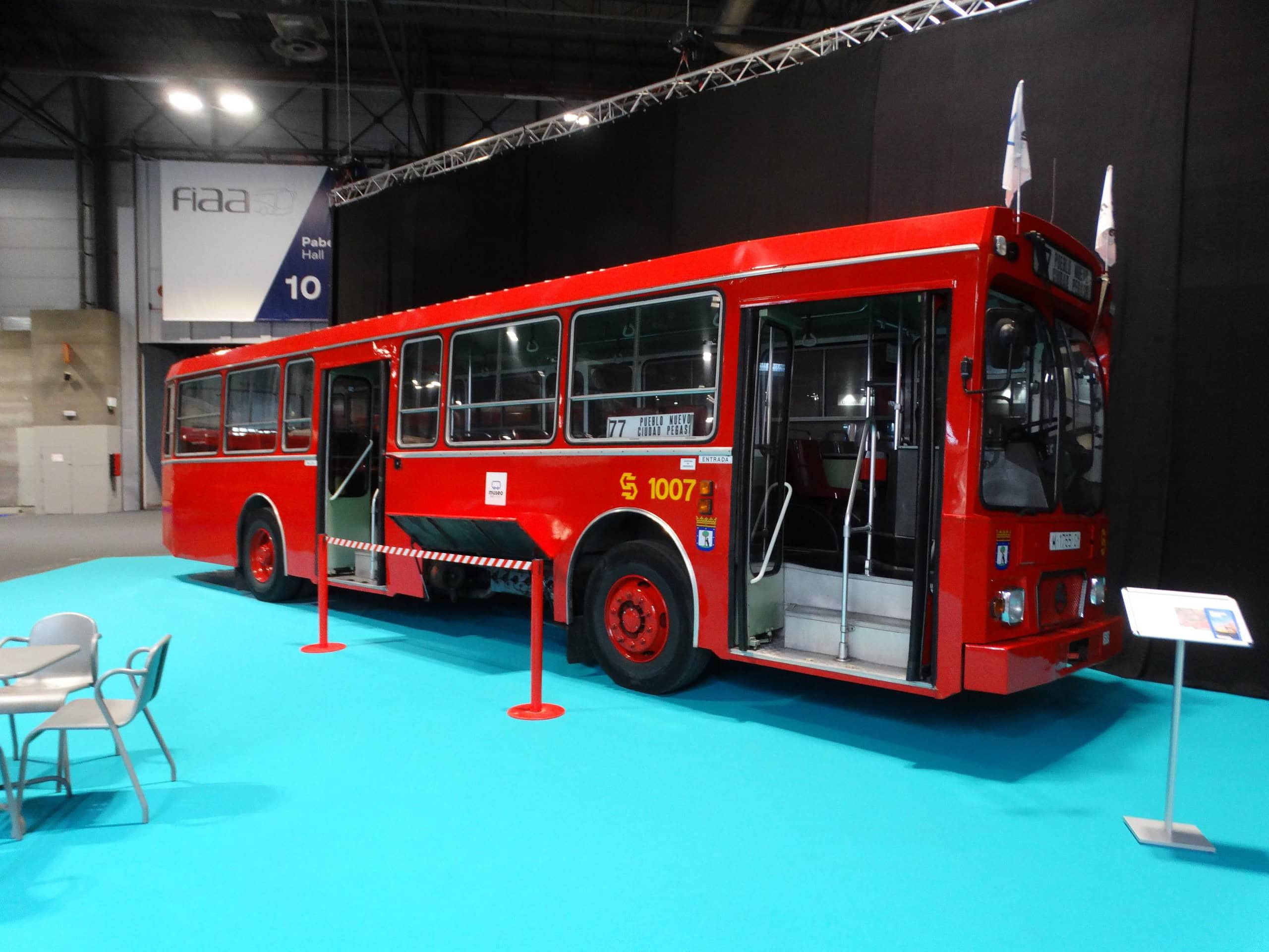 Un autobús rojo antiguo, parte de la Feria Internacional del Autobús y del Autocar, se exhibe en el interior sobre un piso turquesa, captando la atención de los visitantes.