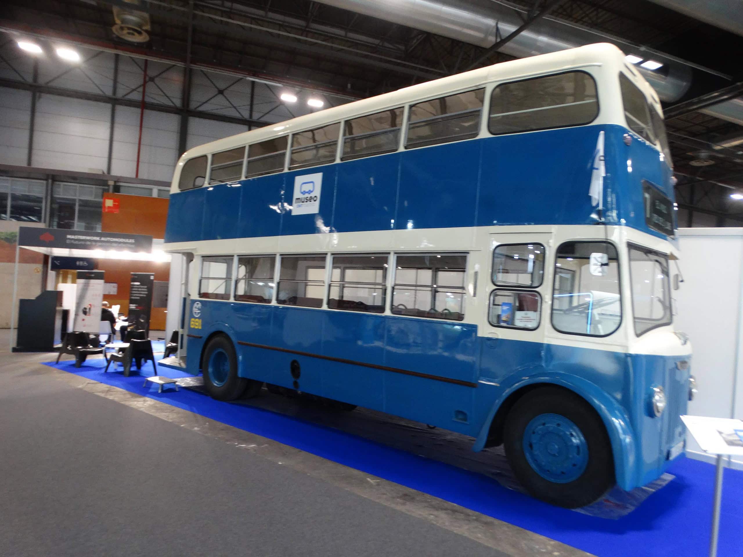 Un autobús antiguo de dos pisos de color azul y crema se exhibe en el interior de la Feria Internacional del Autobús y del Autocar, con puestos de información cercanos.