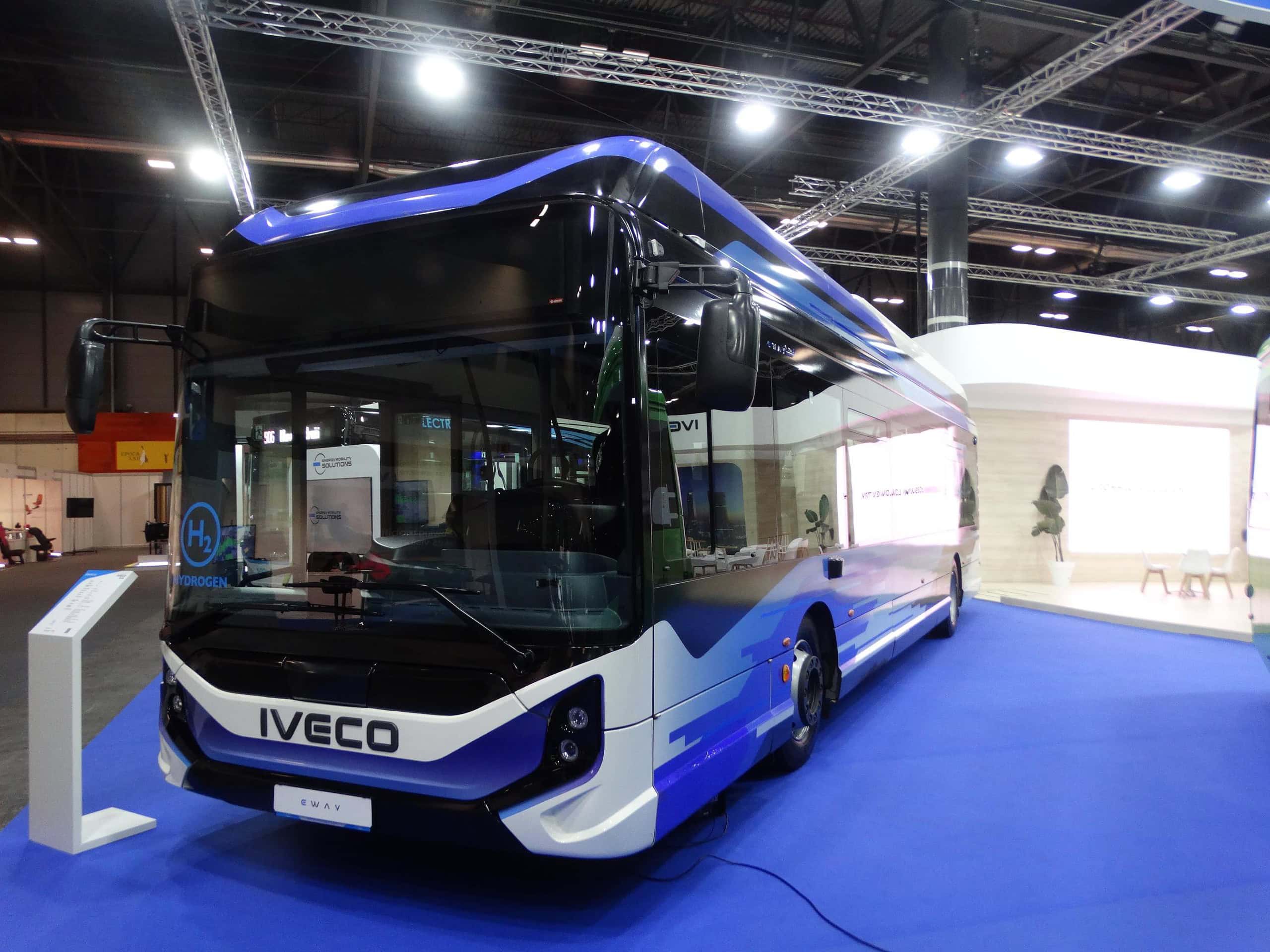 Un elegante autobús Iveco azul y blanco expuesto en la Feria Internacional del Autobús, captando la atención de los visitantes mientras recorren la exposición interior alfombrada de azul.