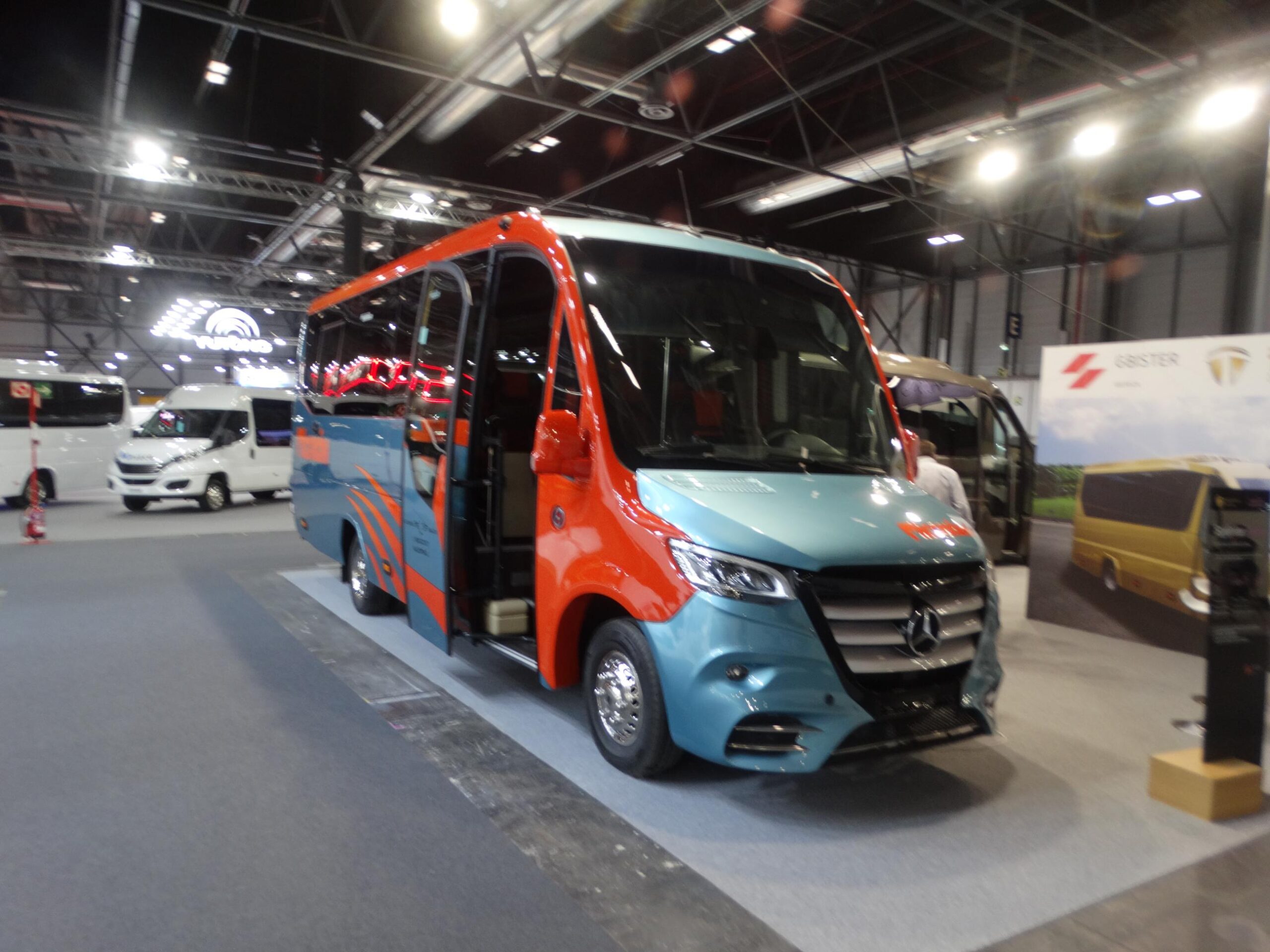 Un colorido autobús de la Feria Internacional del Autobús se exhibe en el interior, iluminado por luces superiores, con una serie de otros vehículos, incluido un elegante Autocar, en el fondo.