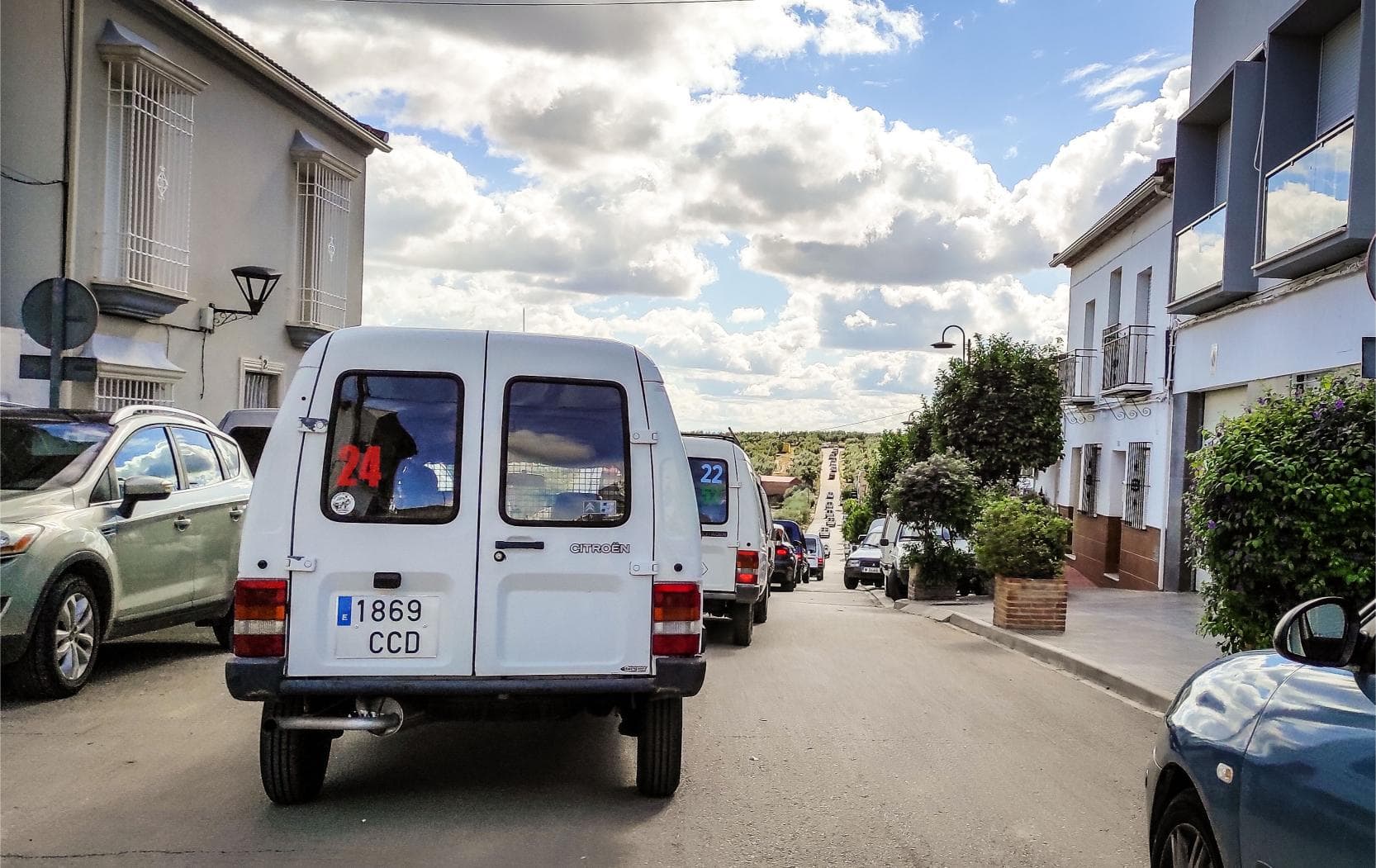 Un clásico Citroën C-15 se desliza por una calle suburbana, enclavado entre autos estacionados y bajo un cielo nublado, insinuando aventuras como la Expedición Camper-15 a Marruecos.