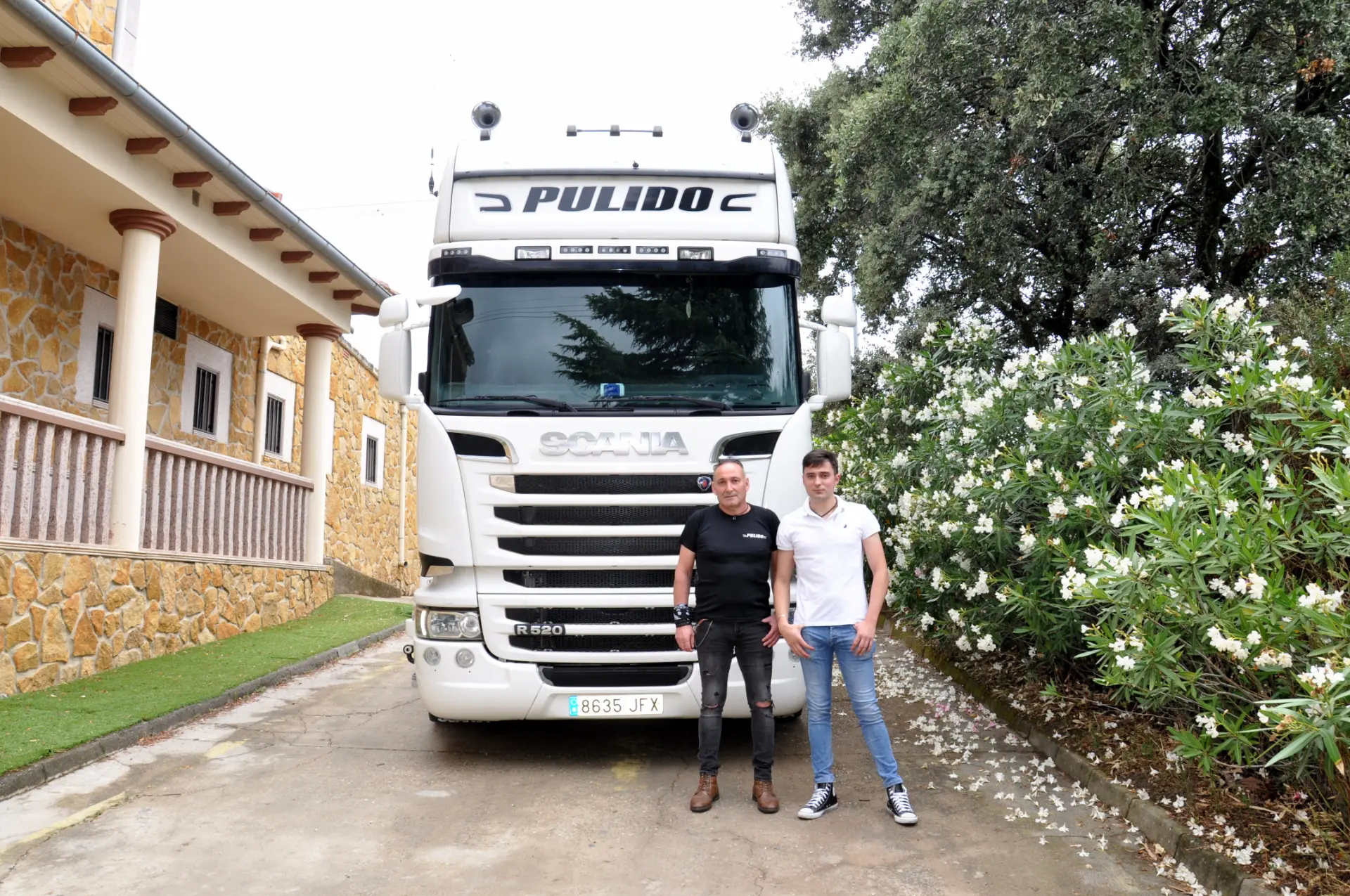 Dos personas están paradas frente a un gran camión blanco en una entrada, con flores y un edificio al fondo, como un boceto automático viviente que captura los momentos espontáneos de la vida.