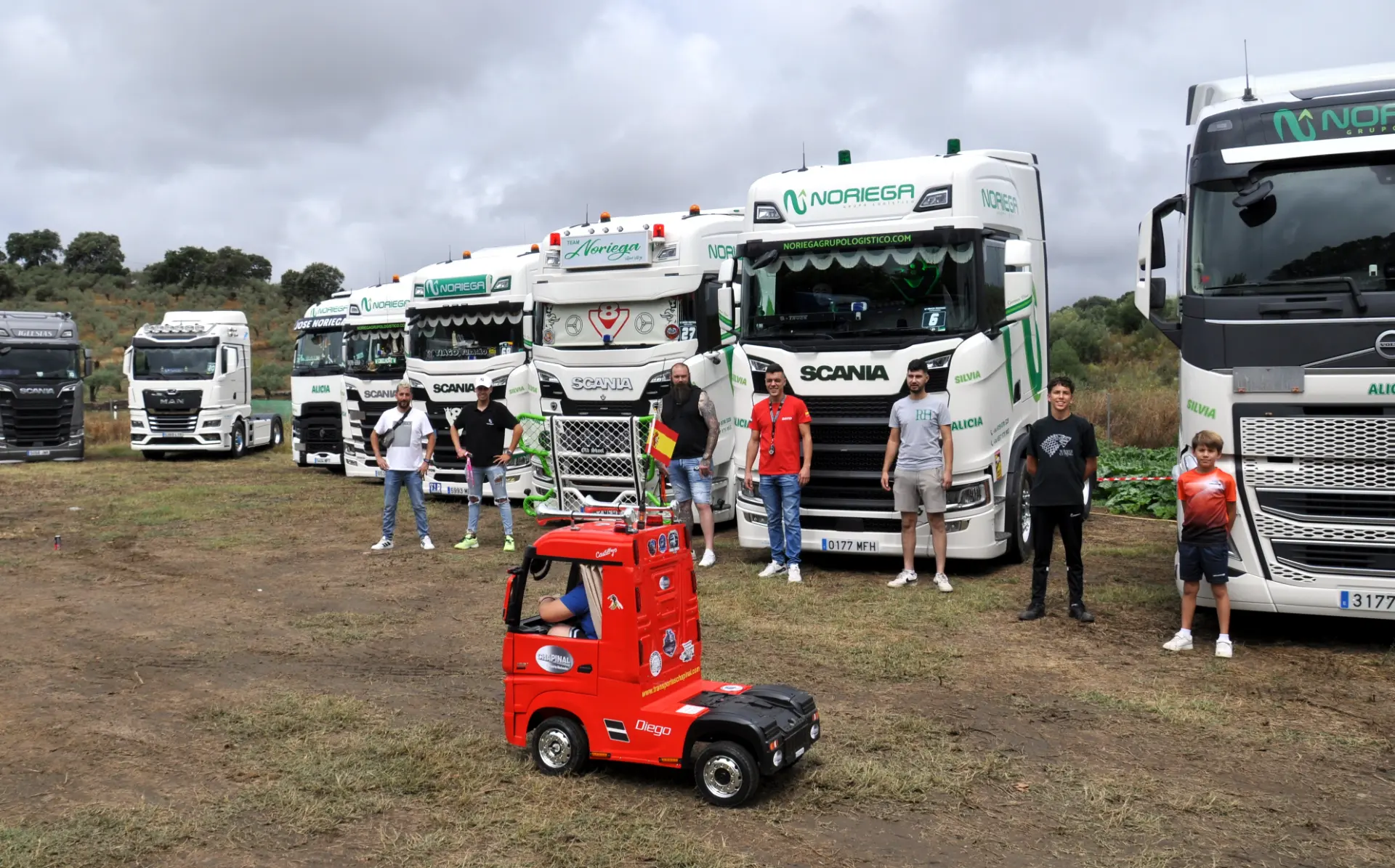 Una hilera de camiones estacionados sobre el césped, con gente de pie junto a ellos, crea una escena animada. En primer plano se encuentra un pequeño camión de juguete rojo, aparentemente ajeno al bullicio que lo rodea: un borrador automático perfecto en esta vívida instantánea.
