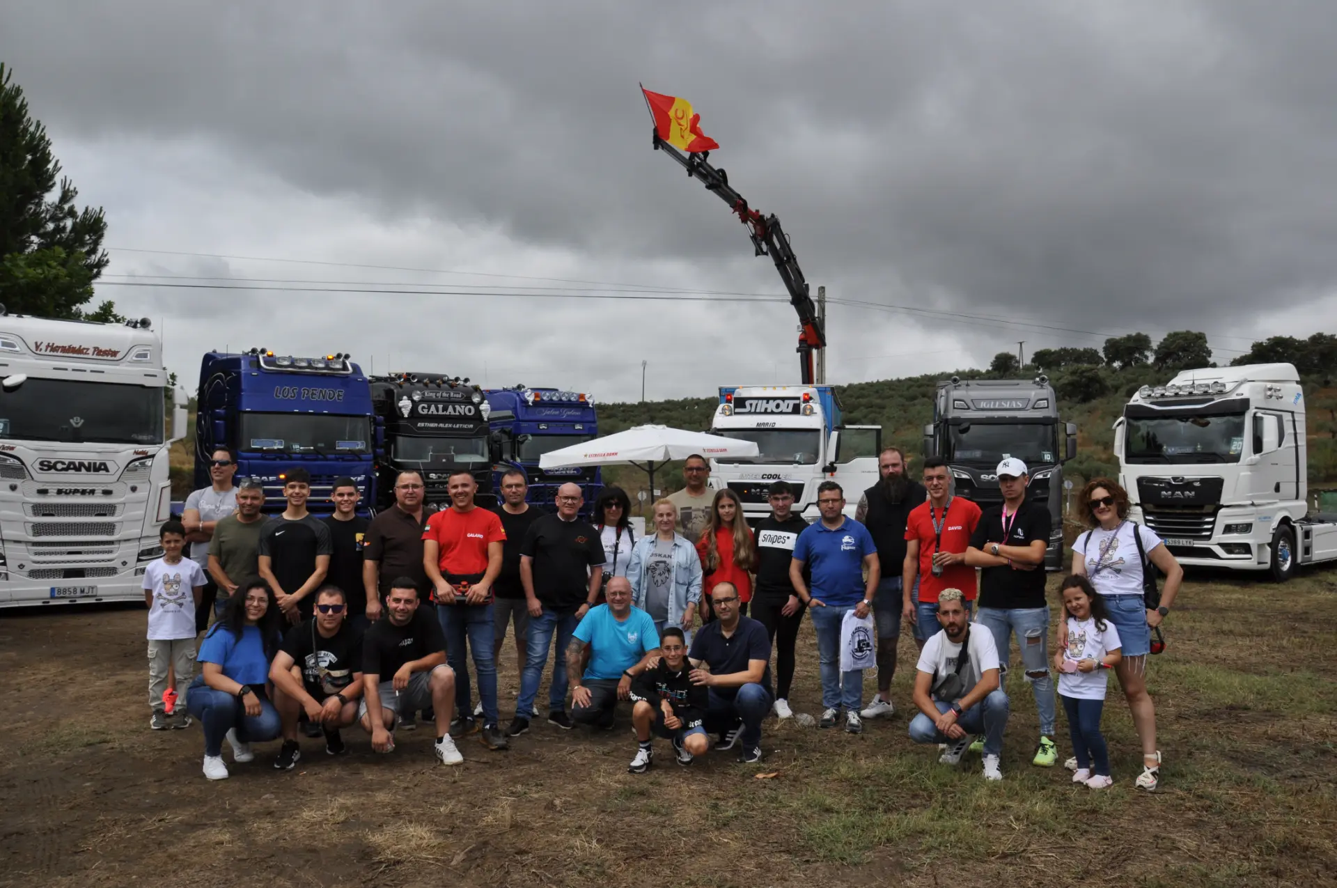 Un grupo de personas posa orgullosamente frente a camiones bajo un cielo nublado, con una bandera izada en una grúa como fondo, capturando el momento como un borrador automático.