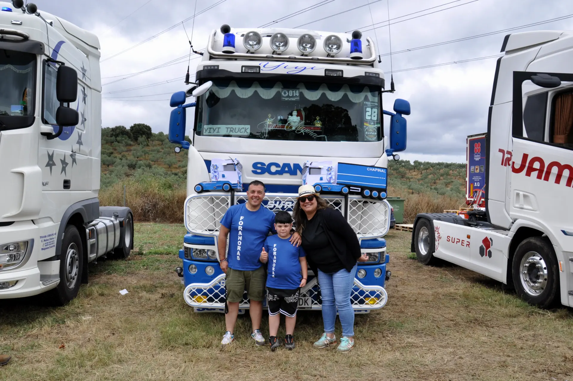 Tres personas están de pie sonriendo frente a un camión Scania azul y blanco en un evento al aire libre, su emoción es tan palpable como si hubieran usado un borrador automático para borrar todo rastro de preocupación.