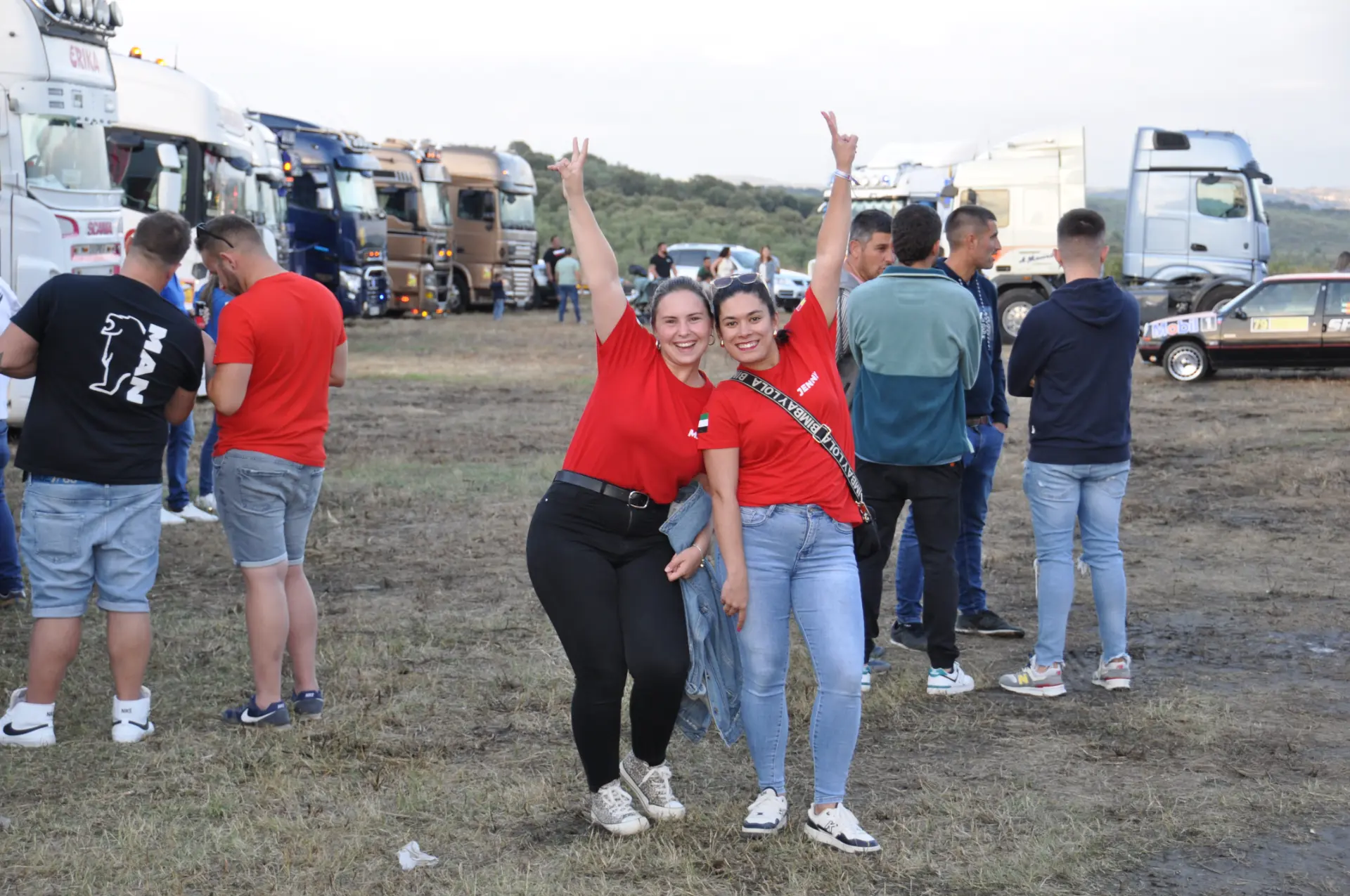 Dos personas con camisas rojas posan felices con símbolos de paz en una reunión de camiones al aire libre, abrazando el espíritu del borrador automático.