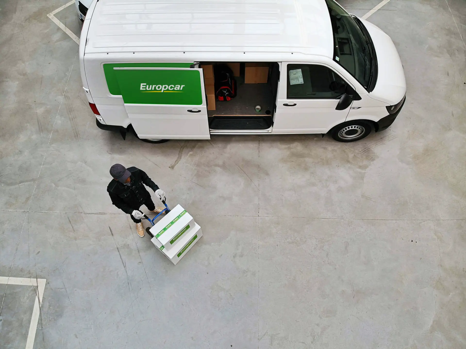 Una persona con cajas pasa junto a una furgoneta Europcar verde brillante en un concurrido aparcamiento, captada desde arriba.