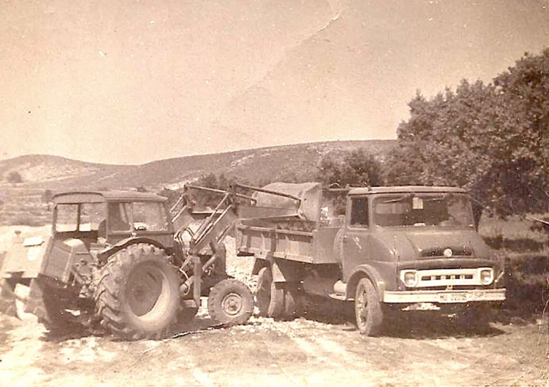 Fotografía vintage de un tractor y un camión en un campo rural con colinas y árboles al fondo, capturando la esencia del campo de Sevilla.