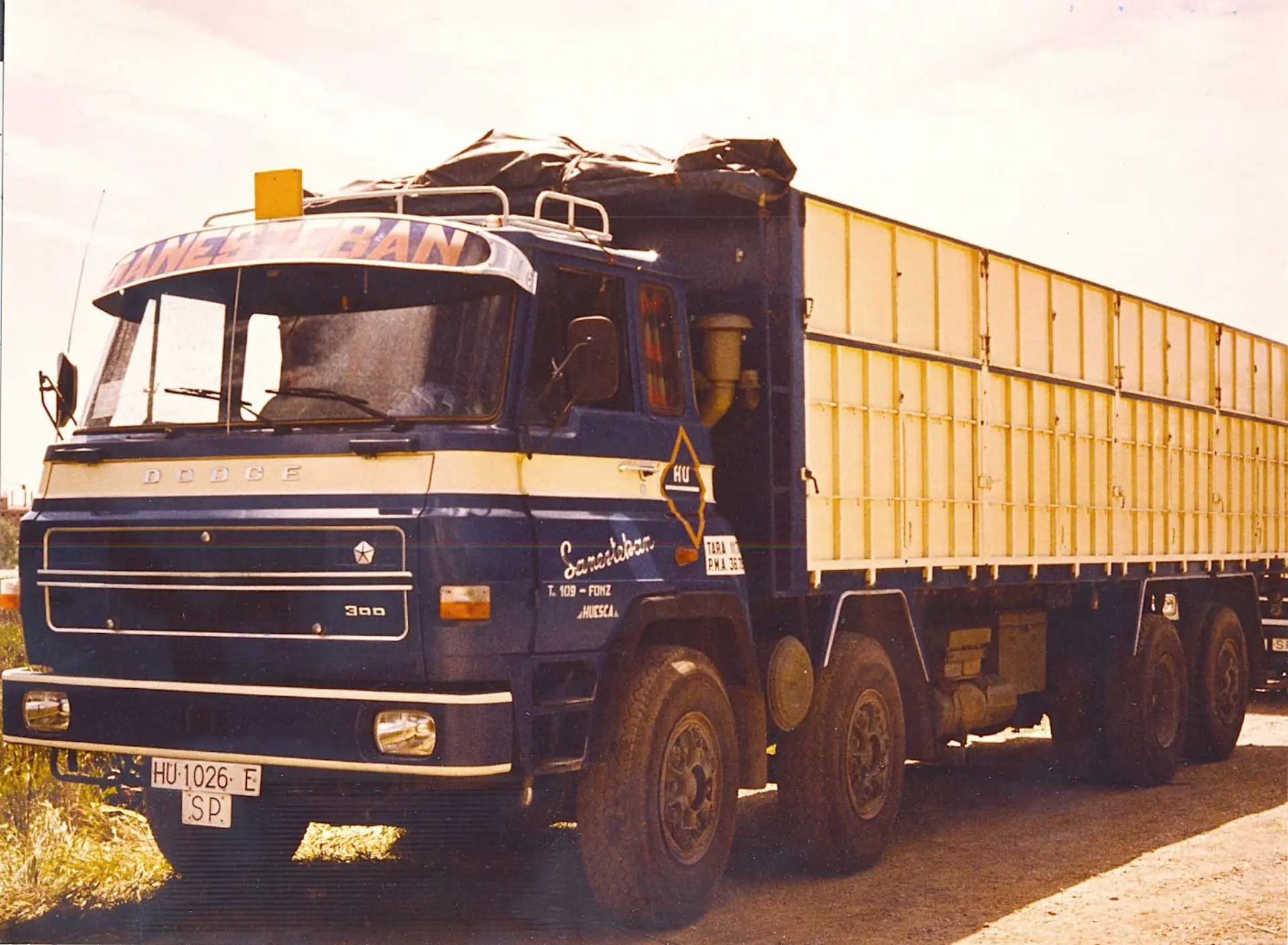 Un camión Dodge azul de época con un gran contenedor de carga amarillo, que recuerda los viajes de Antonio Plata por Sevilla, está estacionado al aire libre. Con sus distintivos COCTRAMO, este vehículo clásico cuenta una historia de viajes y comercio a cielo abierto.