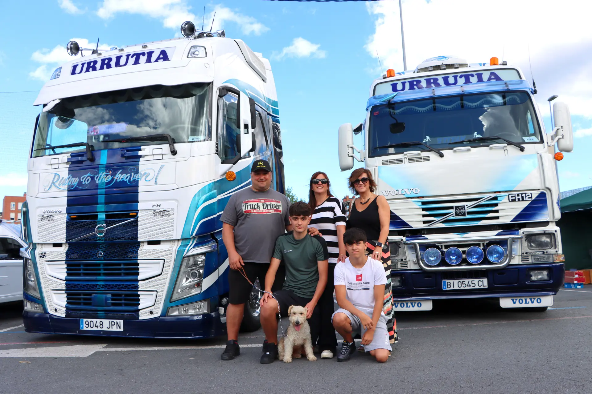 Una familia con un perro posa alegremente frente a dos camiones Volvo blancos y azules en el animado Festival de Camiones, abrazando el espíritu de "lo que nos une nos reúne".