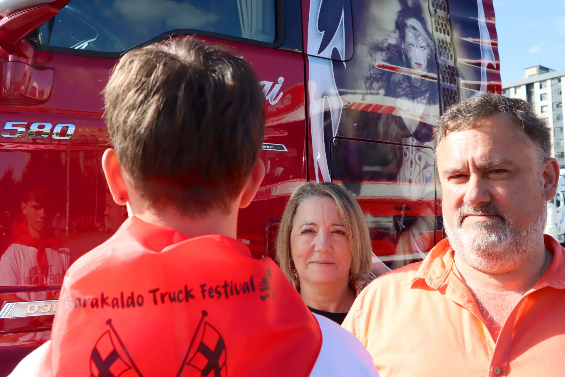 Tres personas permanecen delante de un camión rojo decorado en el vibrante Festival del Camión de Barakaldo, donde el espíritu de nos une nos reúne une a todos.