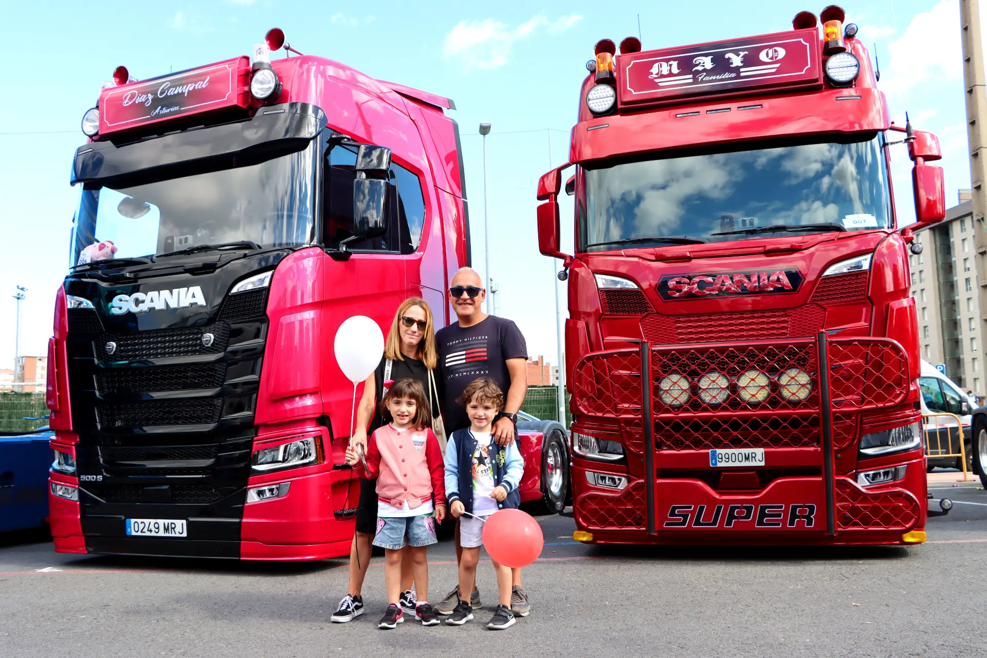 Una familia con globos aerostáticos disfruta del Festival de Camiones de Barakaldo, posando delante de dos camiones Scania rojos brillantes en un evento al aire libre. Son momentos como estos los que verdaderamente encarnan "lo que nos une, nos reúne".