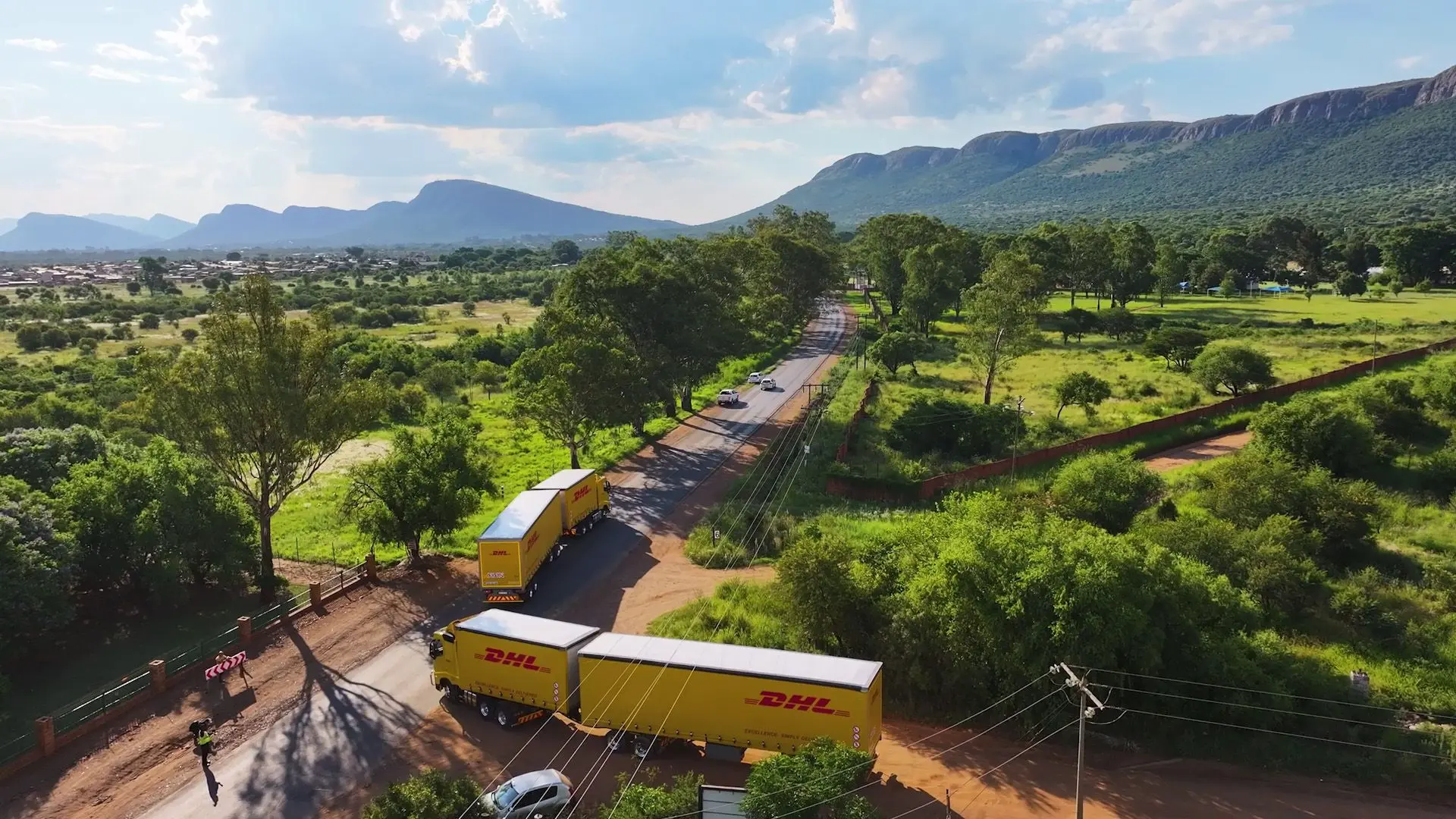 Vista aérea de los camiones de DHL en un exuberante camino rural verde con montañas al fondo, preparados para entregas eficientes en 2024.