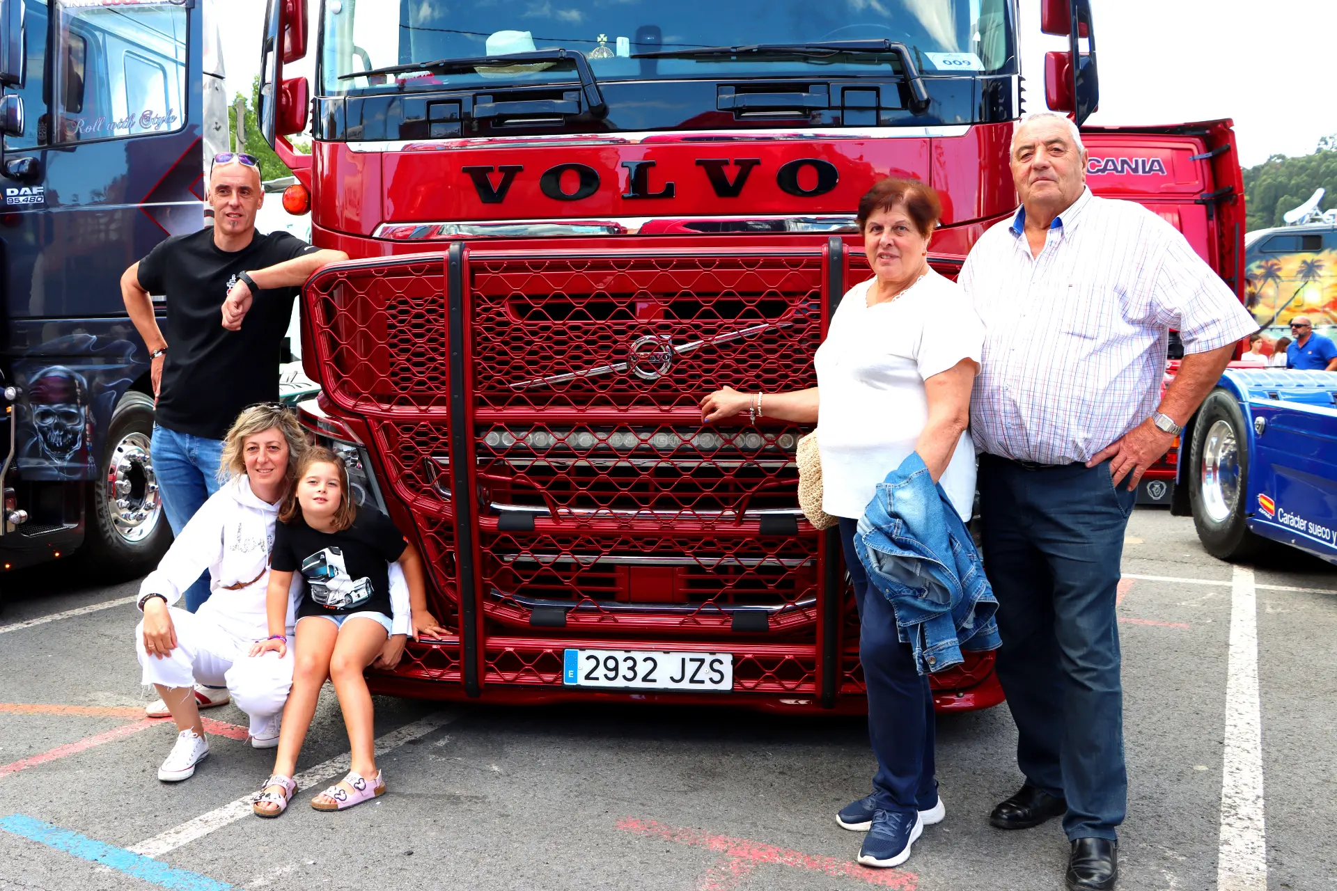 Una familia posa alegremente delante de un gran camión Volvo rojo, sus sonrisas reflejan la alegría que se respira en el Festival de Camiones de Barakaldo en la concurrida zona de aparcamiento.