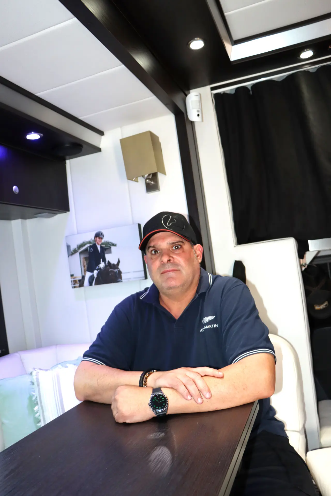 Un hombre está sentado a una mesa en una habitación con poca luz y cortinas oscuras; la atmósfera recuerda al Festival de Camiones de Barakaldo. Detrás de él, una foto en la pared captura recuerdos que nos unen y unen a las personas a través de experiencias compartidas.
