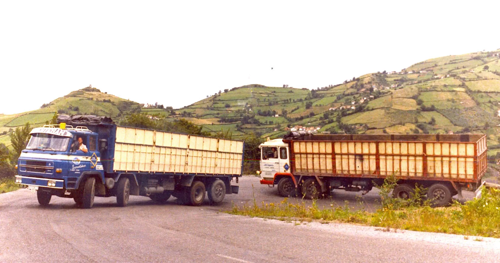 Dos grandes camiones COCTRAMO avanzan por una carretera rural con verdes colinas al fondo, que recuerdan las rutas panorámicas cercanas a Sevilla.