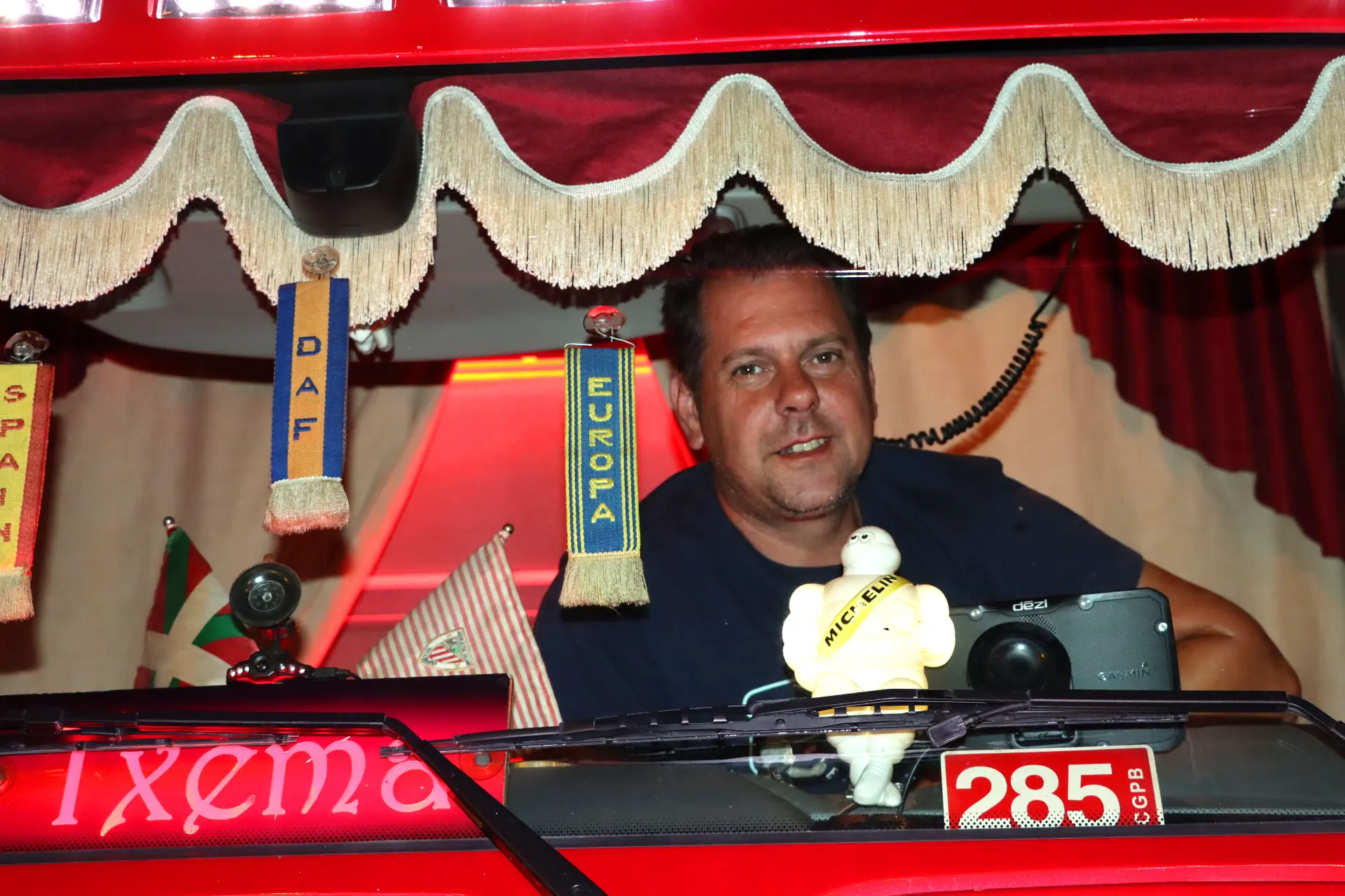 Un hombre dentro de una cabina de camión roja adornada con banderas y una figura de Michelin sonríe a la cámara, captando el espíritu festivo del V Barakaldo Truck Festival.