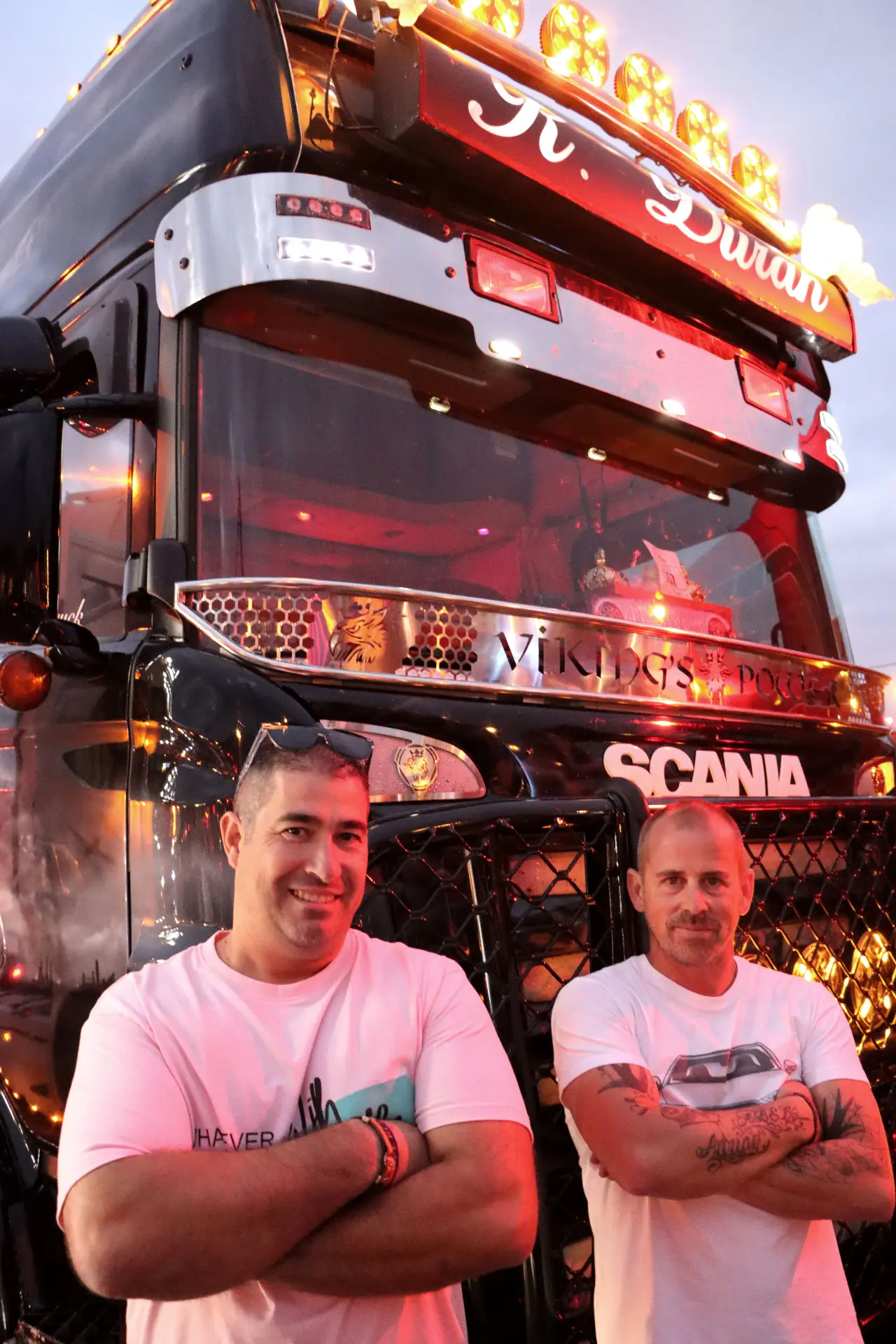 Dos hombres, sonrientes con los brazos cruzados, se encuentran orgullosos frente a un camión Scania decorado al anochecer, capturando el espíritu del Festival de Camiones de Barakaldo.