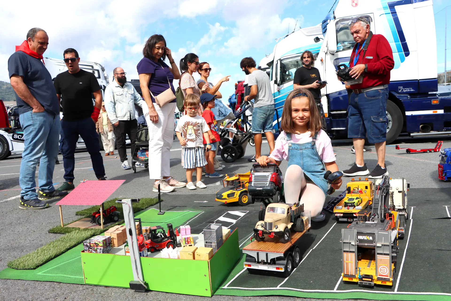 En el Barakaldo Truck Festival, una niña juega alegremente con camiones de juguete sobre una alfombra callejera, llamando la atención de adultos y niños allí reunidos. Nos reunimos en un momento de simple diversión, mostrando lo que nos une.