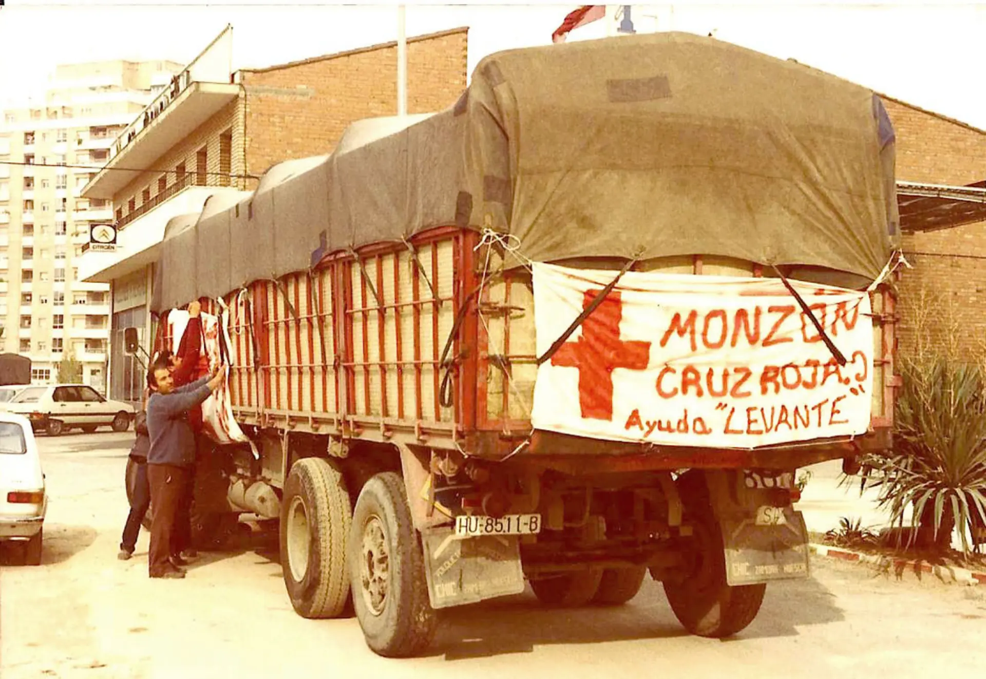 Un camión con una carga envuelta en una lona y con una pancarta con la leyenda "Cruz Roja" está estacionado junto a un edificio en Sevilla. Cerca de allí, Antonio Plata supervisa la operación con precisión alineada a los estándares logísticos de COCTRAMO.