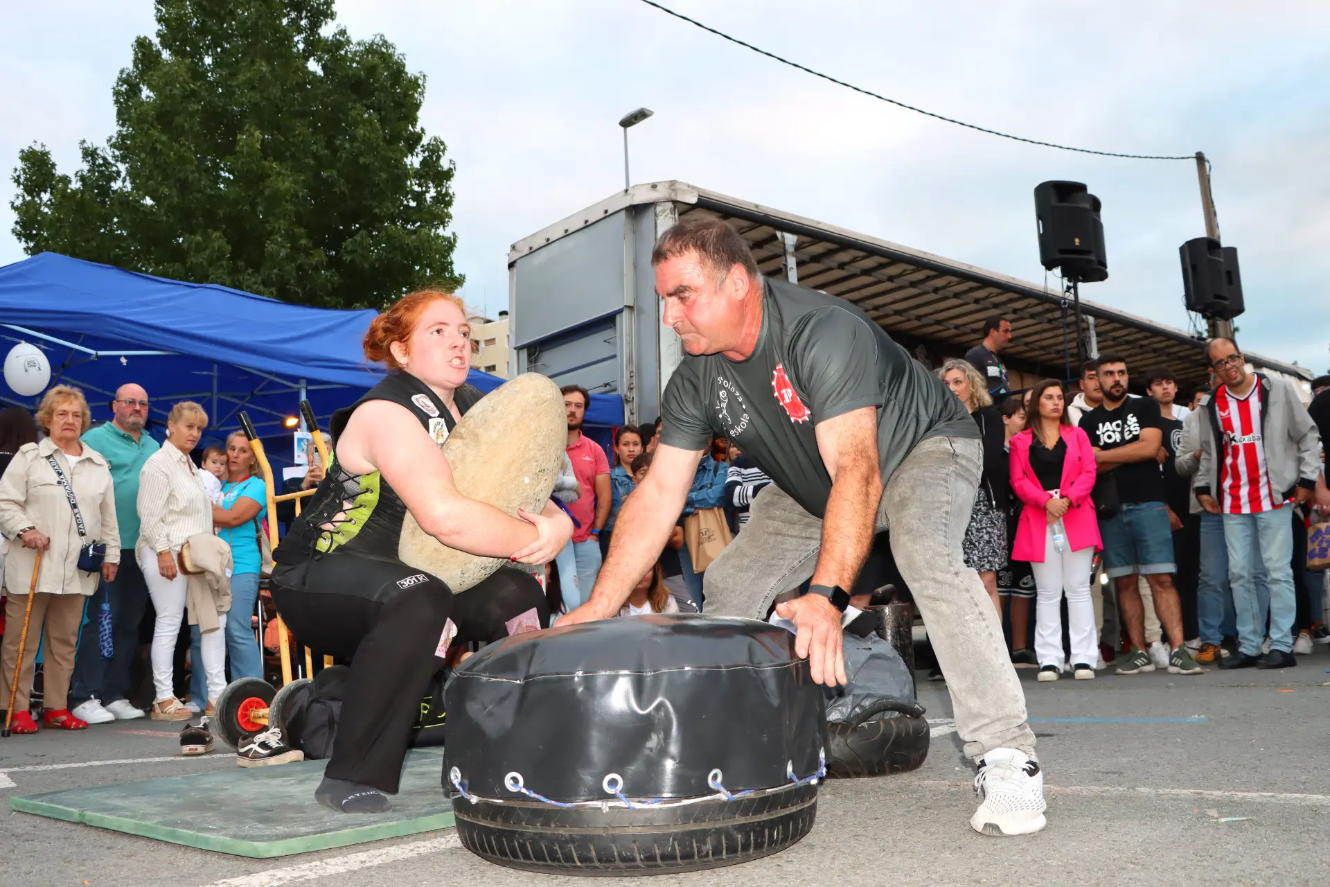 Una mujer y un hombre compiten en una prueba de strongman, levantando pesas en el Barakaldo Truck Festival, con una multitud entusiasta observando de fondo.