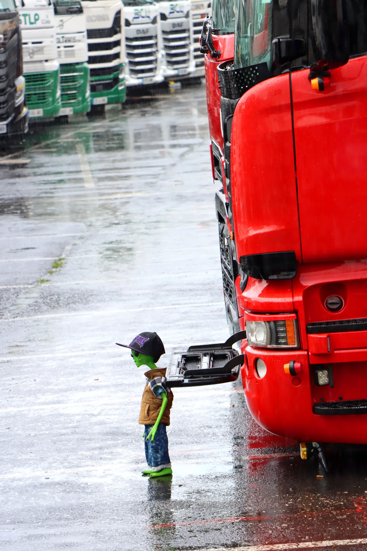 Un niño con gorra y chaqueta permanece bajo la lluvia junto a un gran camión rojo en el animado Festival de Camiones de Barakaldo, sin que el clima afecte su entusiasmo.