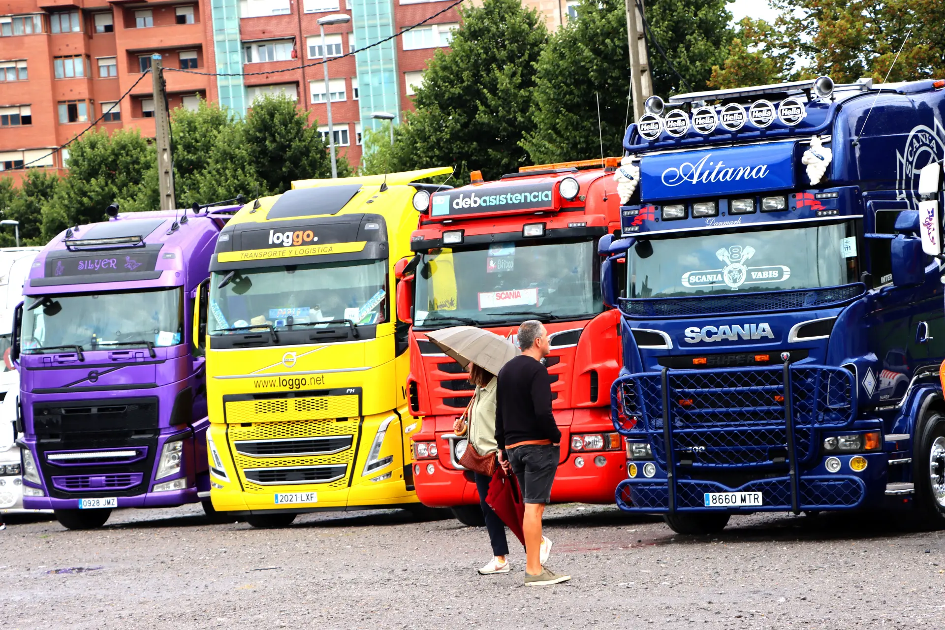 Varios camiones coloridos se alinean en un vibrante desfile mientras la gente pasea y disfruta del animado ambiente del Festival de Camiones de Barakaldo. De fondo, edificios encantadores y árboles frondosos crean un entorno pintoresco para esta gran celebración de los gigantes del motor y el espíritu comunitario.