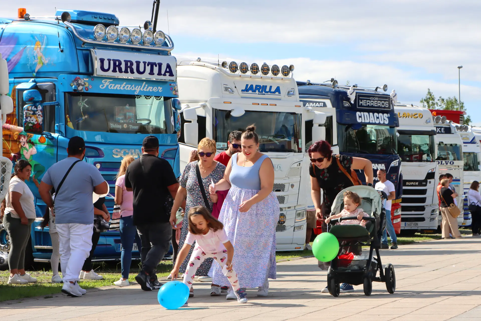 Las familias disfrutan de un día al aire libre en el Festival de Camiones de Barakaldo, donde se aparcan en filas camiones de colores. Los niños se divierten jugando con globos bajo el cielo soleado, lo que contribuye a la atmósfera festiva.