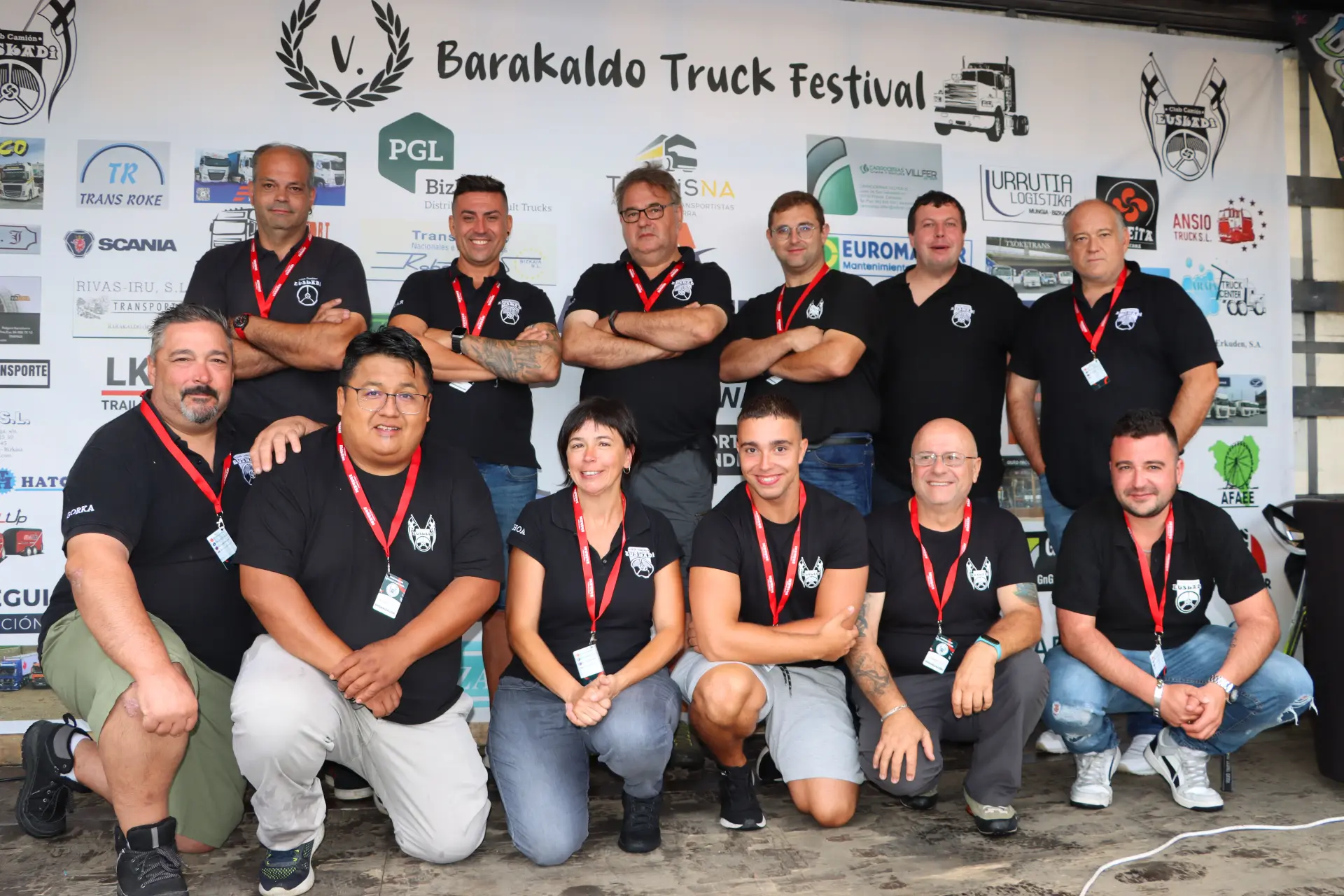Un grupo de personas con camisetas negras iguales en el Festival de Camiones de Barakaldo, abrazando el espíritu de "lo que nos une", posan orgullosamente delante de pancartas de patrocinadores.