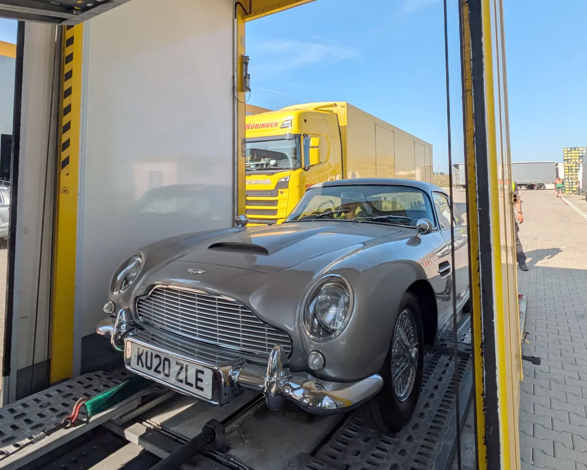 Un automóvil clásico plateado se encuentra en un camión de transporte, con un vehículo DHL amarillo vibrante en el fondo, estacionado en una zona industrial.