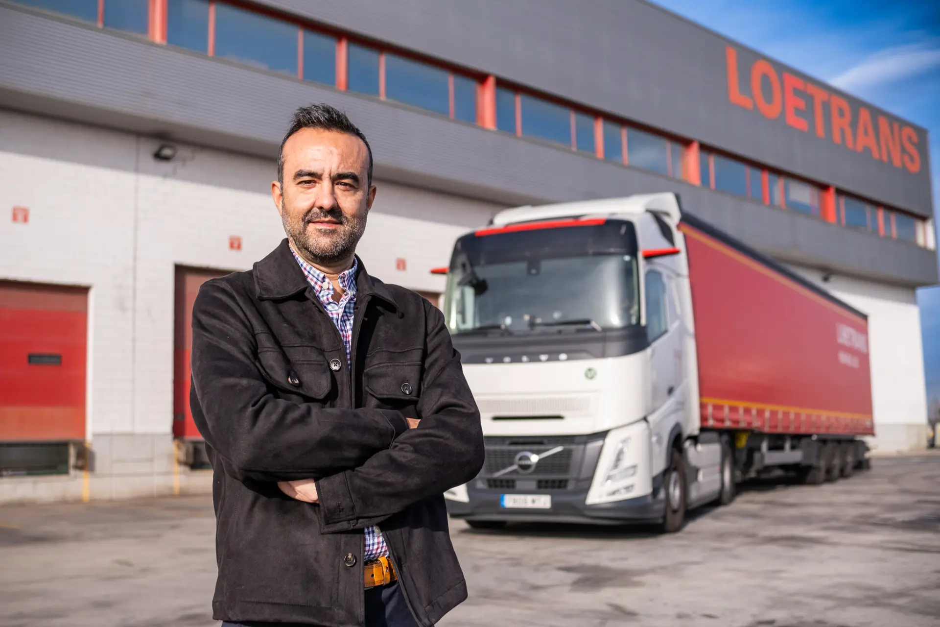 Un hombre con chaqueta se mantiene firme frente a un camión Volvo FH Aero y un edificio con la inscripción "LOETRANS", que simboliza la innovación y el ahorro de combustible.