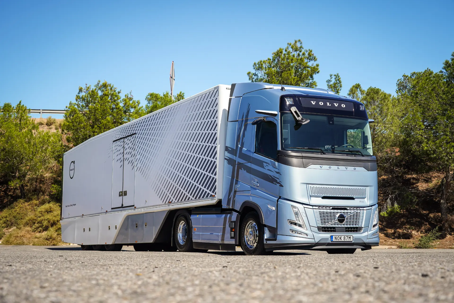Un camión Volvo FH plateado con remolque, equipado con la eficiente tecnología I-Save, está estacionado en un día soleado, rodeado de frondosos árboles.