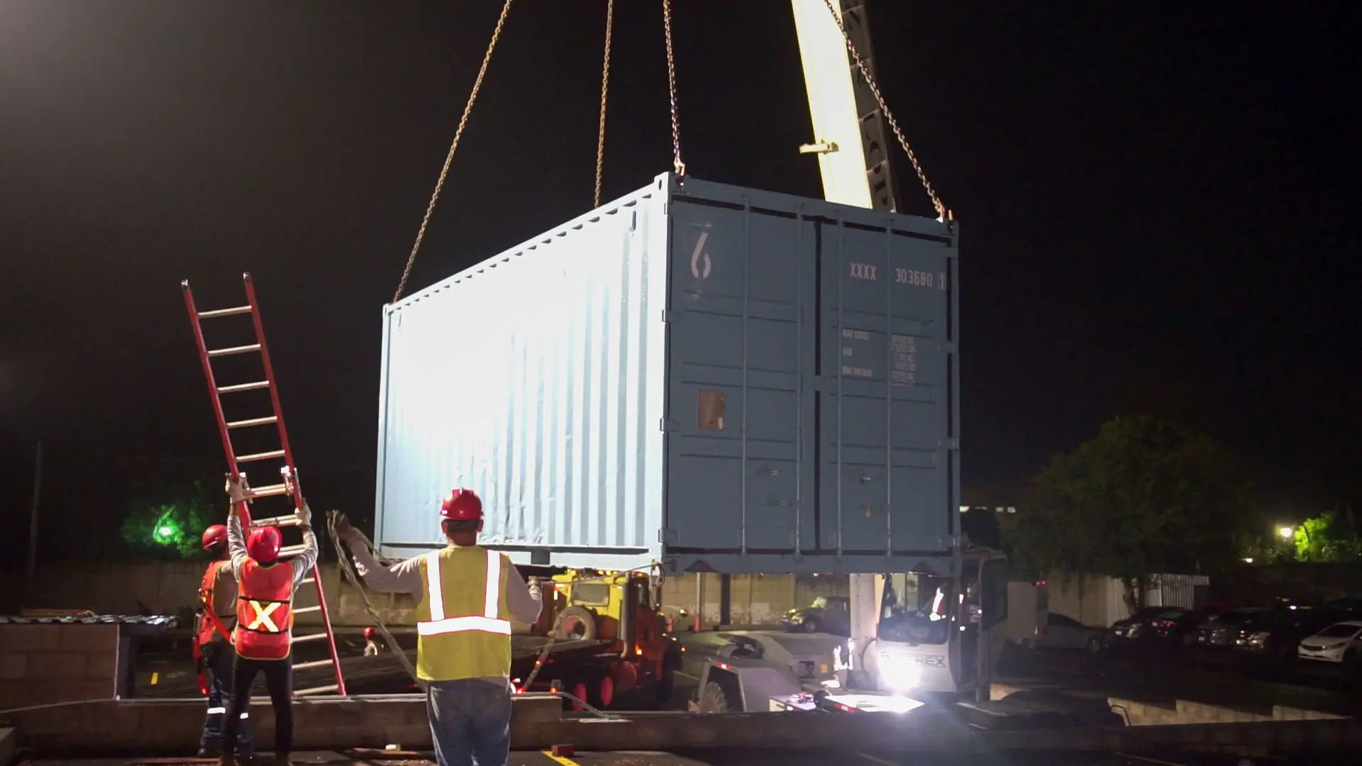 Los trabajadores con equipo de seguridad guían un contenedor de carga, marcado con el logotipo de DHL, mientras es elevado por una grúa bajo el cielo nocturno. Esta escena prepara el escenario para 2024, que promete entregas espectaculares las 24 horas del día.