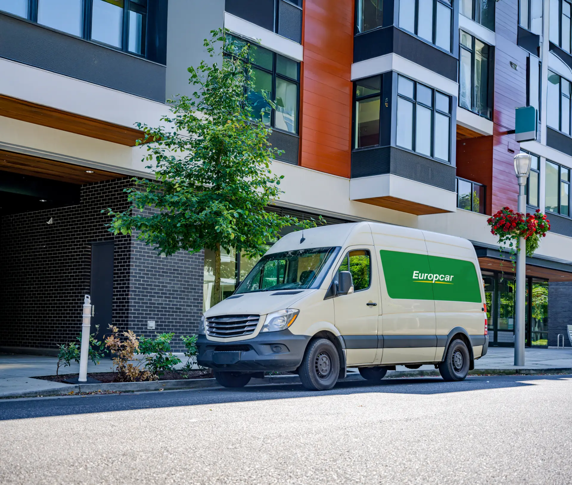 Una elegante furgoneta Europcar está estacionada en una calle frente a un edificio moderno, rodeada de frondosos árboles y coloridas cestas de flores colgantes.