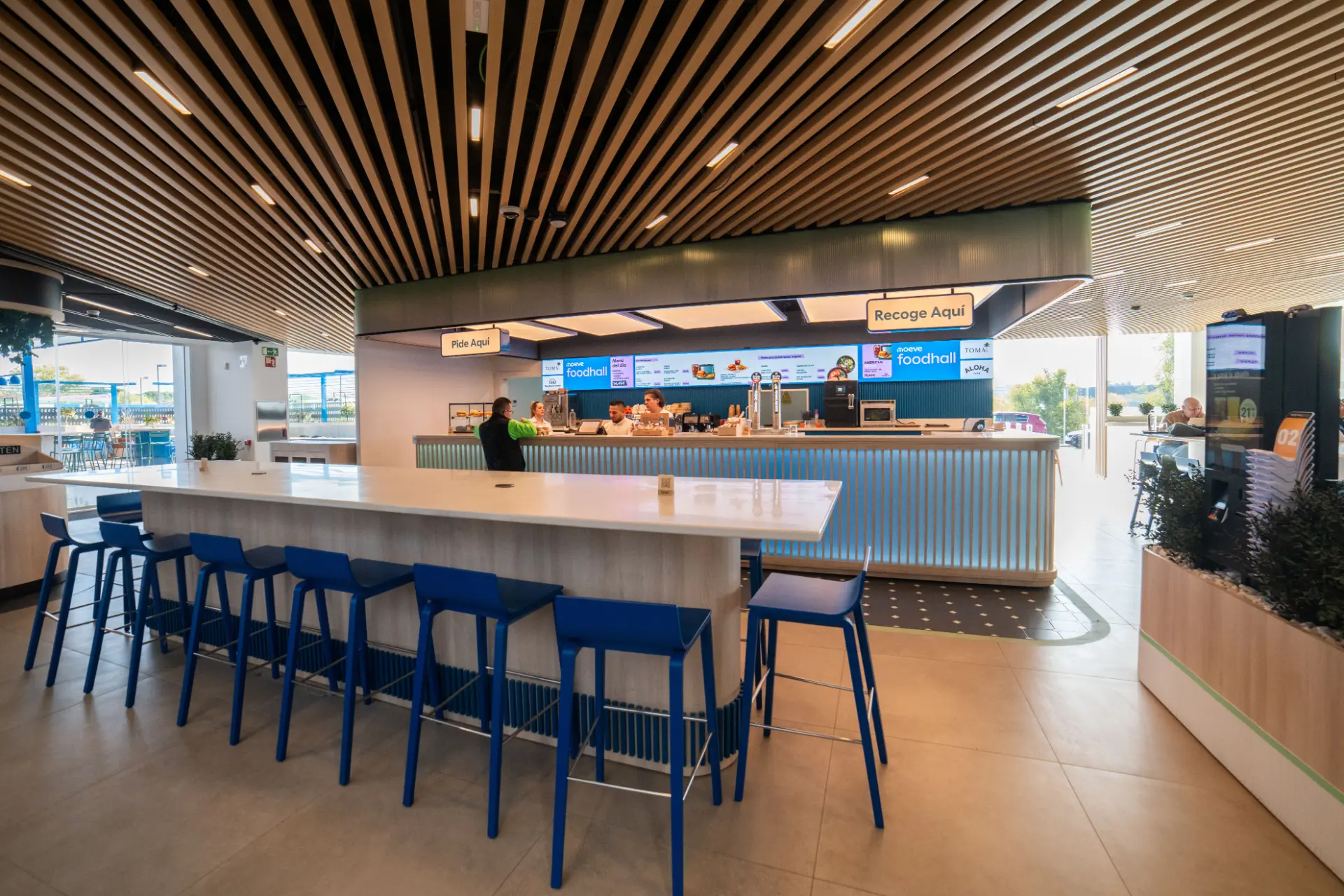 Revoluciona tu experiencia culinaria en este moderno patio de comidas con una gran mesa y taburetes azules, menús digitales y un interior luminoso y espacioso, perfecto para viajeros en estaciones de servicio.