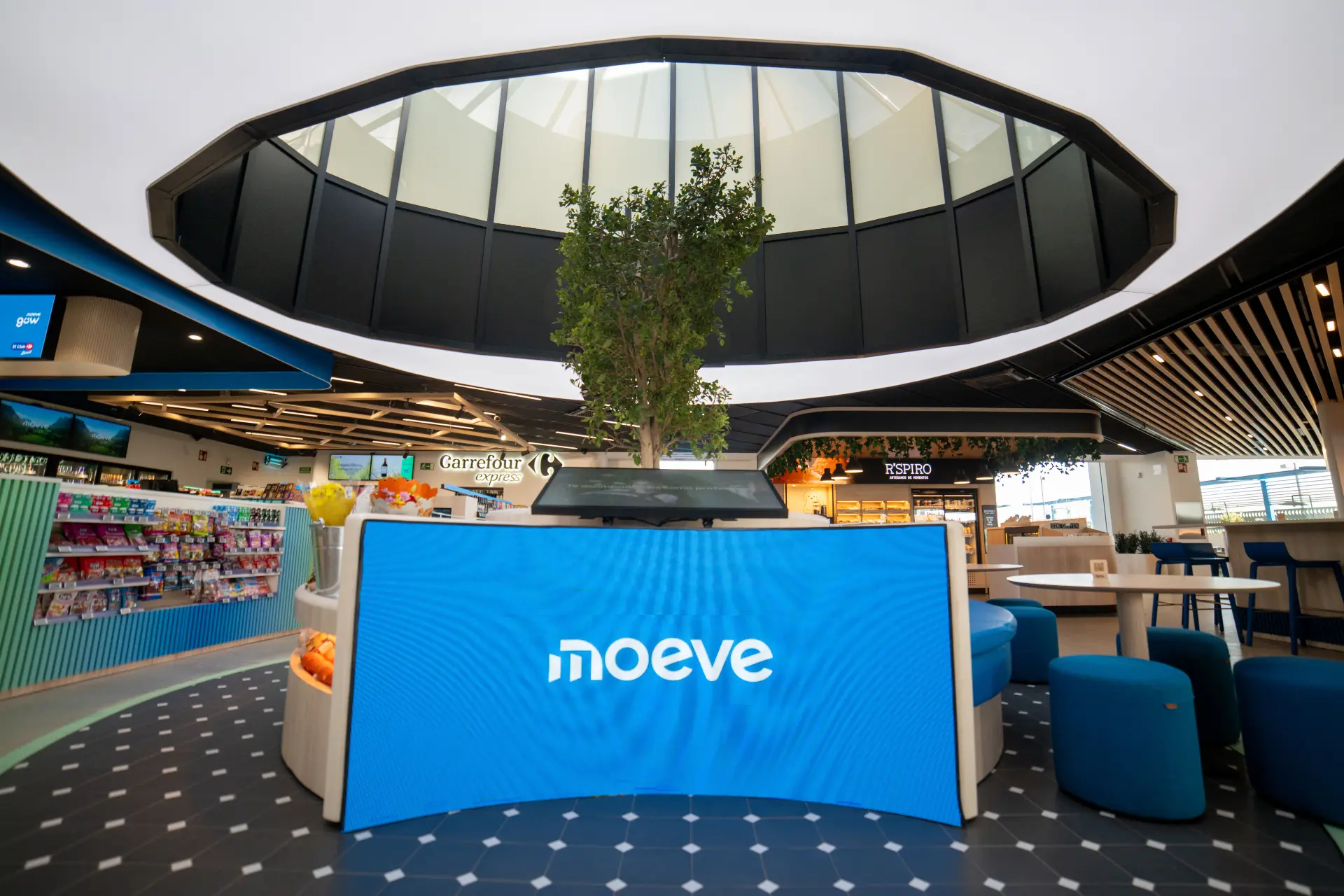 Un interior moderno con un árbol bajo un tragaluz, rodeado de asientos y espacios comerciales, presenta la revolucionaria pantalla azul denominada "moeve", que reinventa las estaciones de servicio.