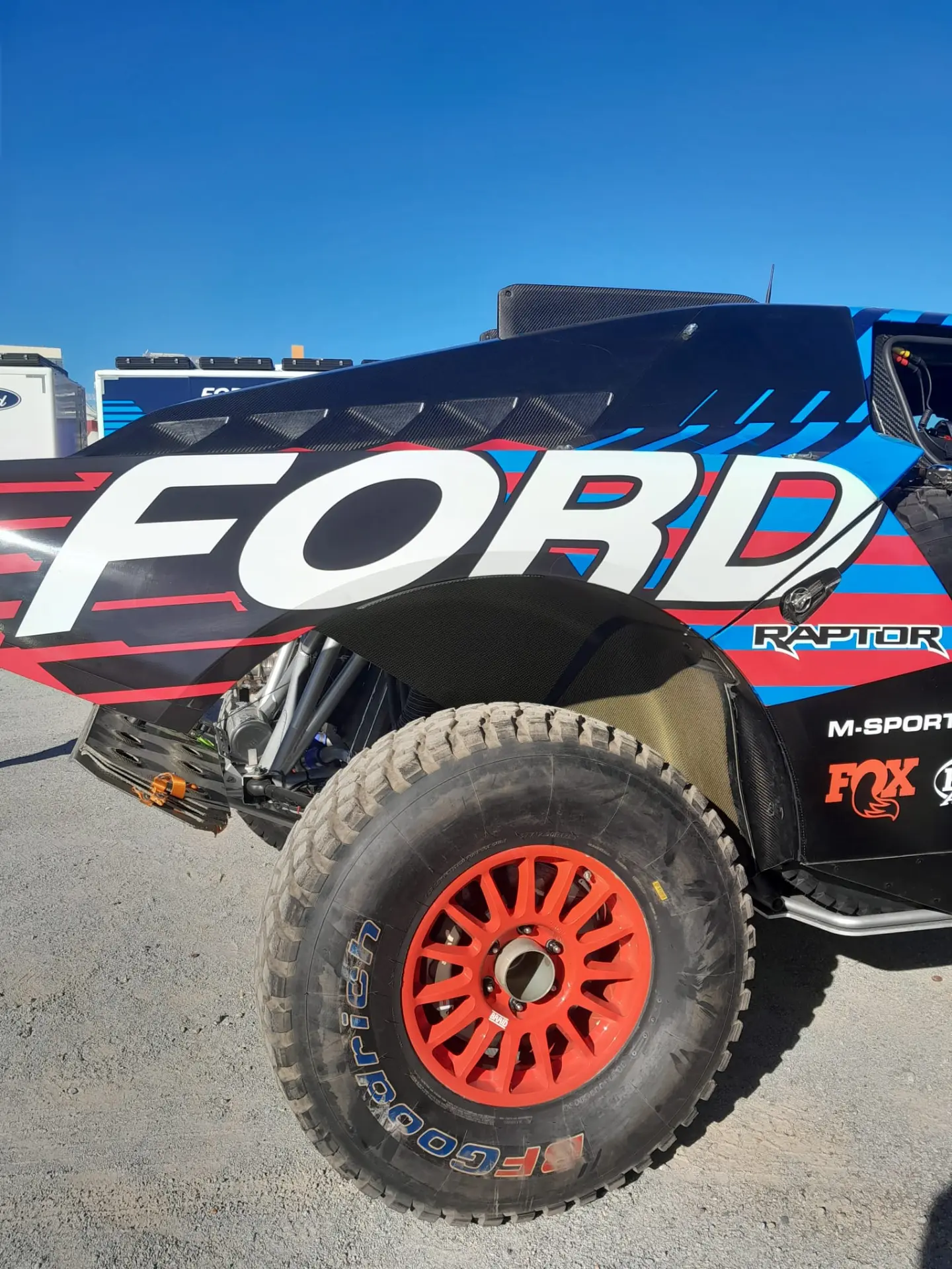 Un primer plano de un camión de carreras Ford Raptor T1+ muestra el estilo característico de Carlos Sainz con ruedas de un rojo vibrante y calcomanías de patrocinadores, todo ello sobre un cielo azul claro. El icónico camión se prepara para el desafío del Dakar 2025.