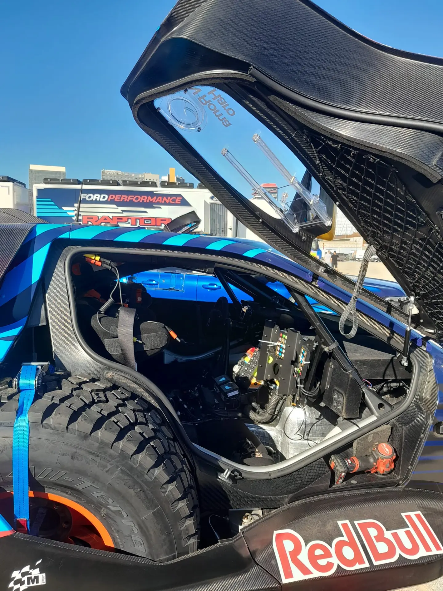 Un capó abierto de un automóvil deportivo revela un interior de alta tecnología con equipamiento de carreras, que recuerda al equipamiento que encontrarías en los vehículos del Dakar 2025, estacionado bajo un cielo azul claro.