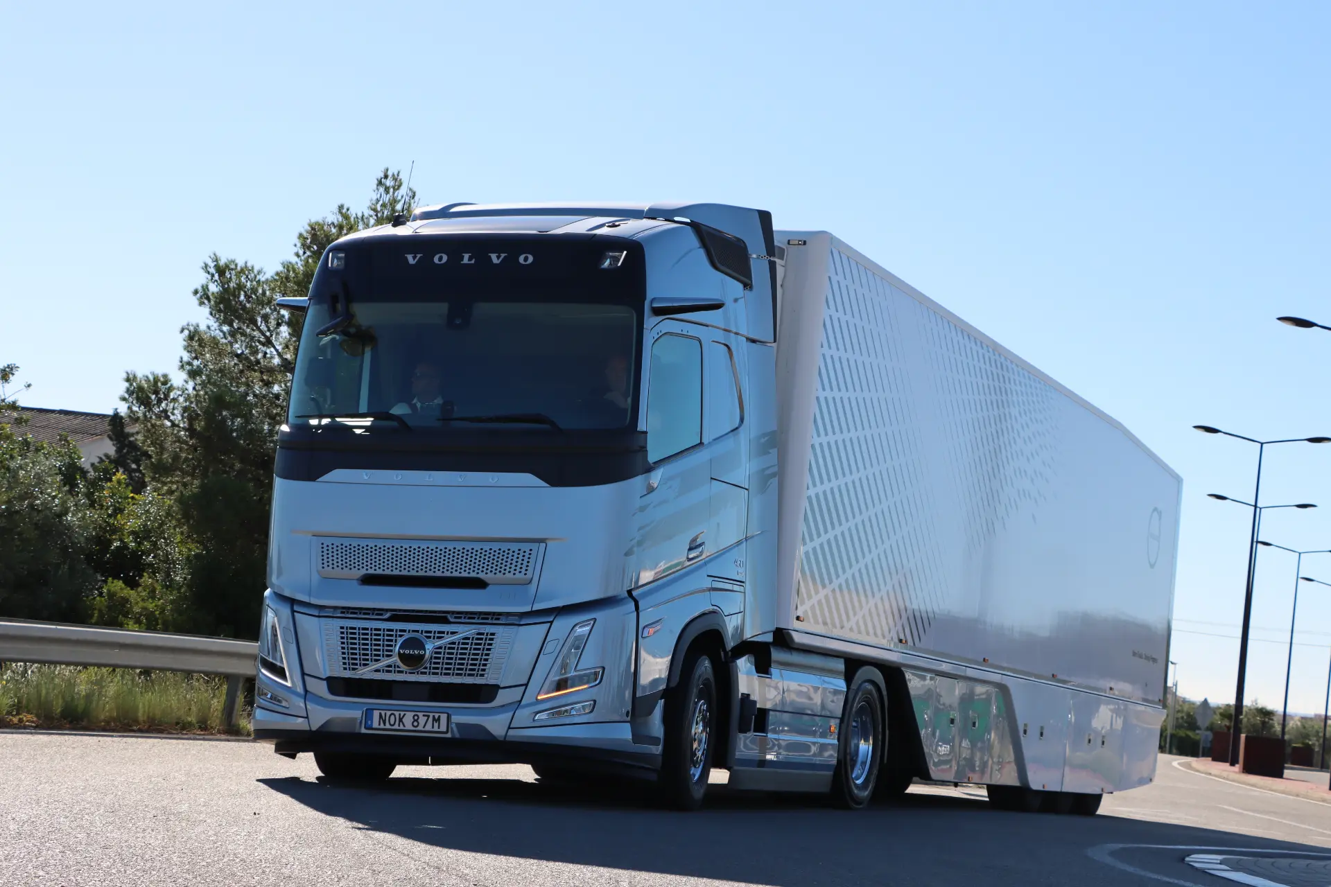 Un camión semirremolque Volvo FH Aero plateado, conocido por su tecnología 460 I-Save, avanza suavemente por una carretera bordeada de árboles e iluminada por farolas.