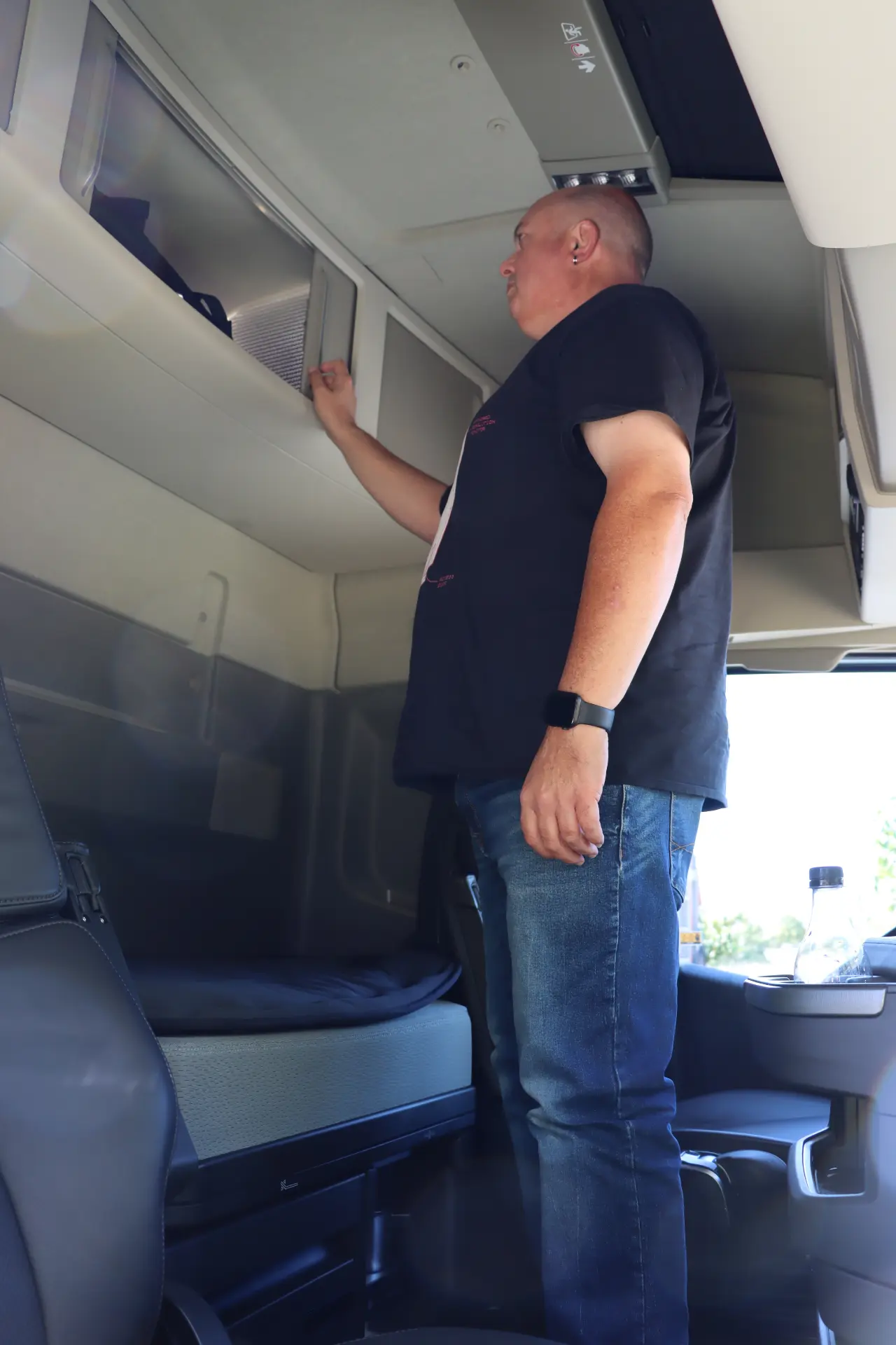 En la cabina de un Volvo FH, un hombre con camiseta negra y vaqueros se acerca al compartimento superior, rodeado por el elegante diseño de la tecnología I-Save.