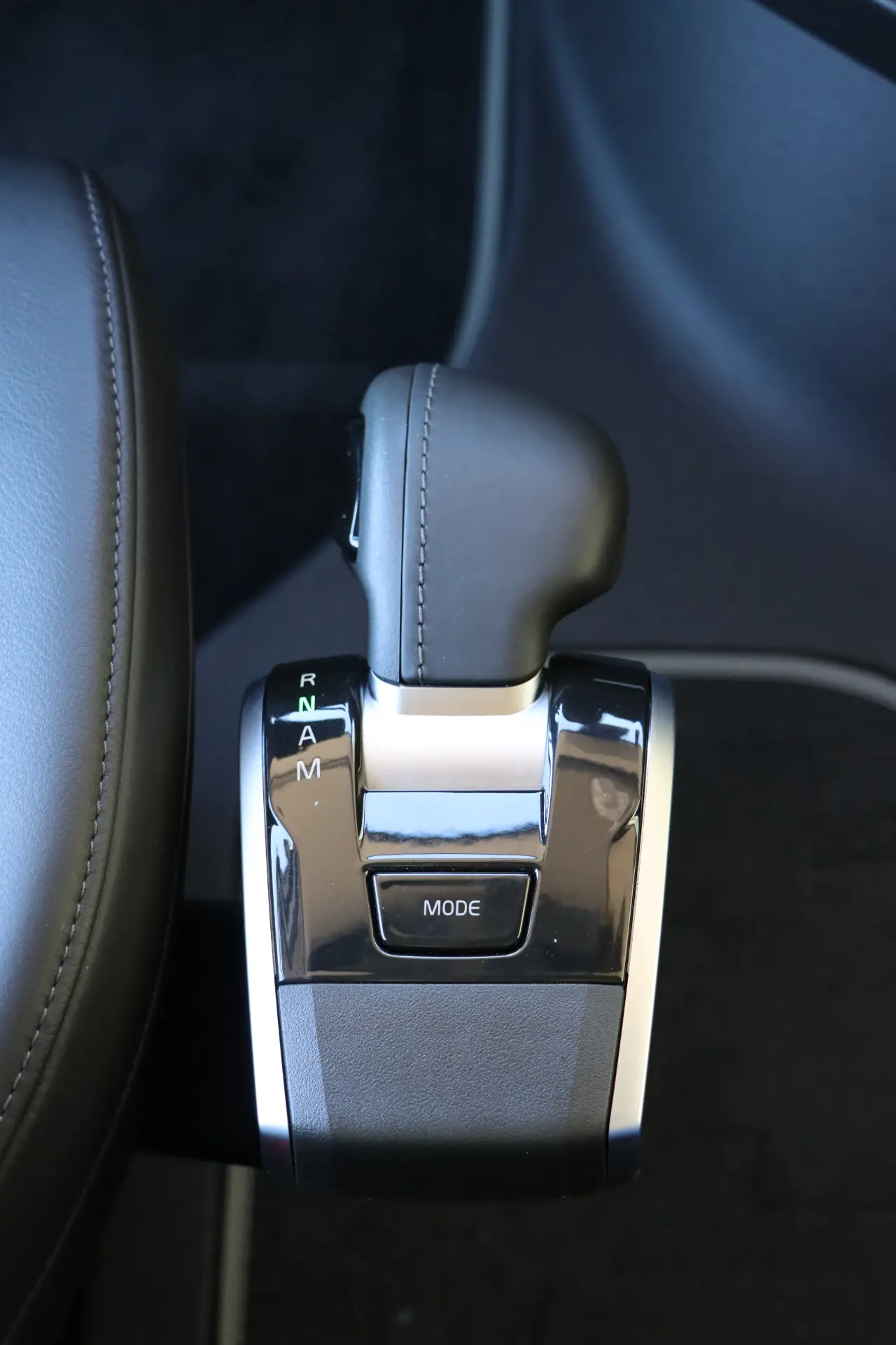 Primer plano de una elegante palanca de cambios de automóvil con opciones P, R, N, D, M y un botón central "MODE" contra una tapicería negra, que se asemeja a la ingeniería de precisión de los modelos Volvo FH o Aero 460 con tecnología I-Save.
