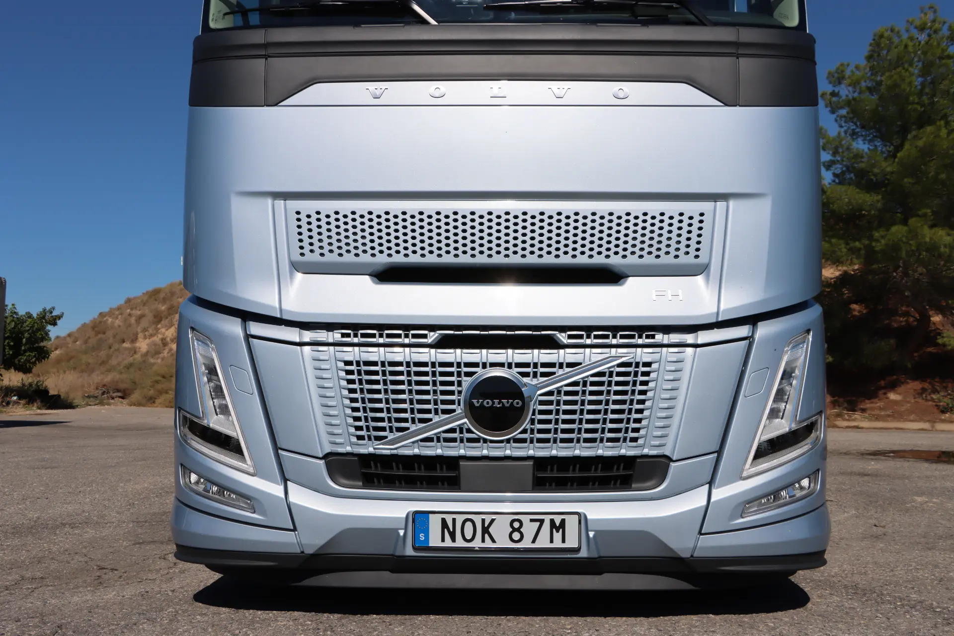 Vista frontal de un camión Volvo FH plateado en un día soleado, con la innovadora tecnología I-Save, estacionado en una superficie pavimentada con colinas y árboles al fondo.