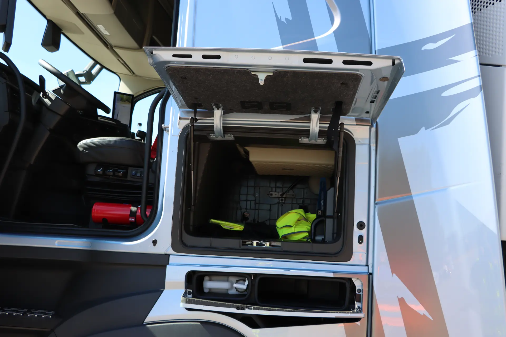 El compartimento de almacenamiento de la cabina del camión Volvo FH Aero está abierto y permite ver el equipamiento de seguridad esencial en su interior. El interior del lado del conductor, diseñado meticulosamente para el modelo 460 I-Save, garantiza tanto funcionalidad como estilo.